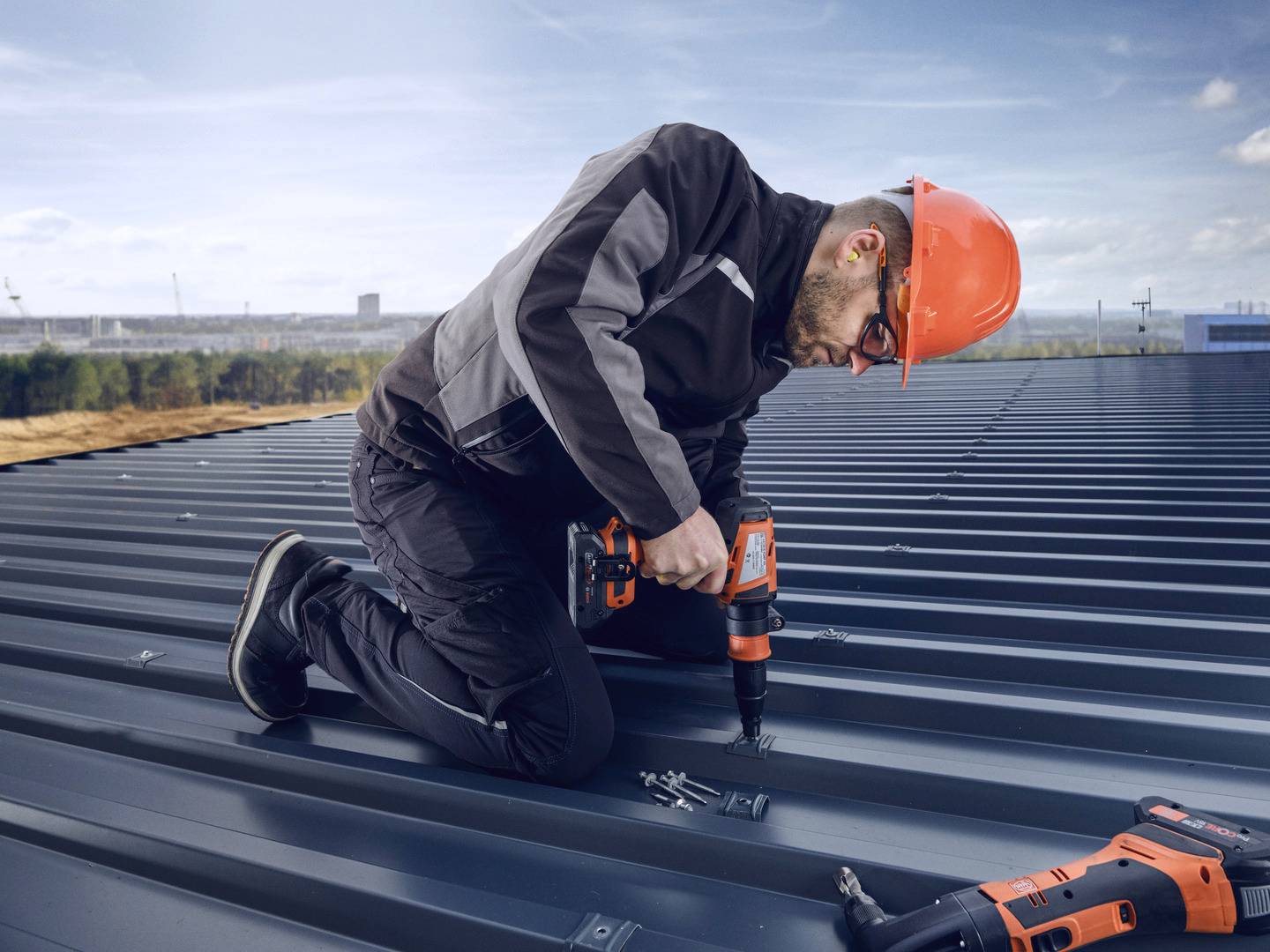 Ein Bauarbeiter mit orangefarbenem Helm befestigt mit einem Akkuschrauber eine Schraube auf einem Blechdach.