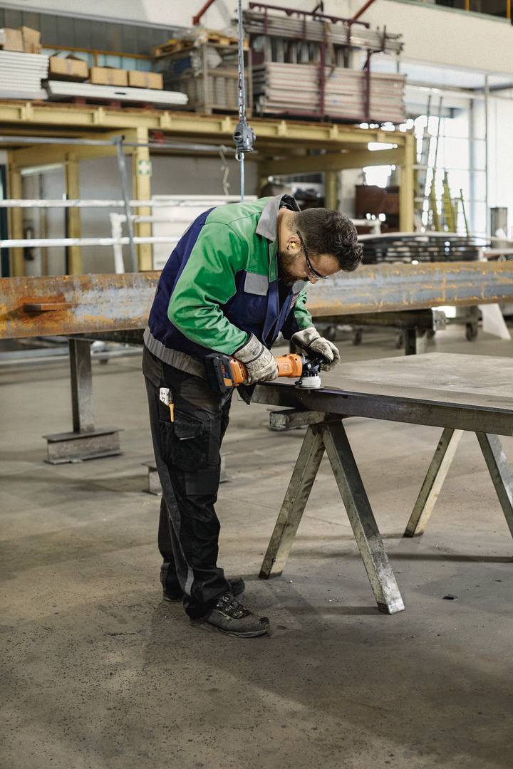 Arbeiter in einer Werkstatt beugt sich über einen Metallbalken, nutzt ein Elektrowerkzeug, um daran zu arbeiten. Umgebung industriell.