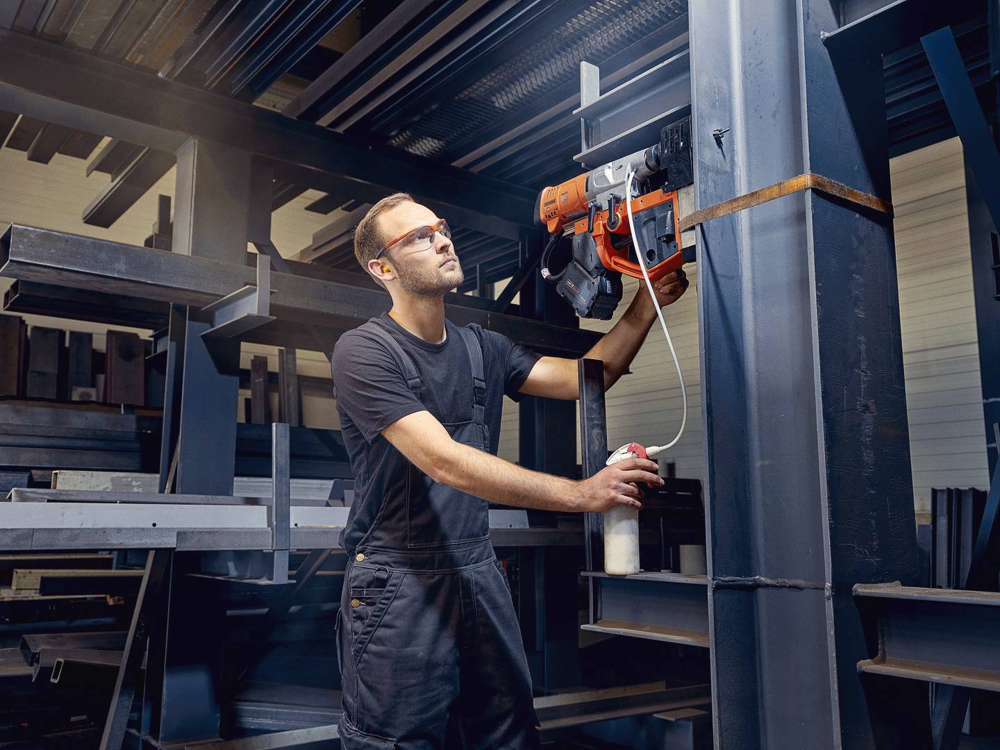 Ein Arbeiter in Schutzbrille und Overall bedient eine elektrische Hebevorrichtung in einer Lagerhalle voller Metallregale.