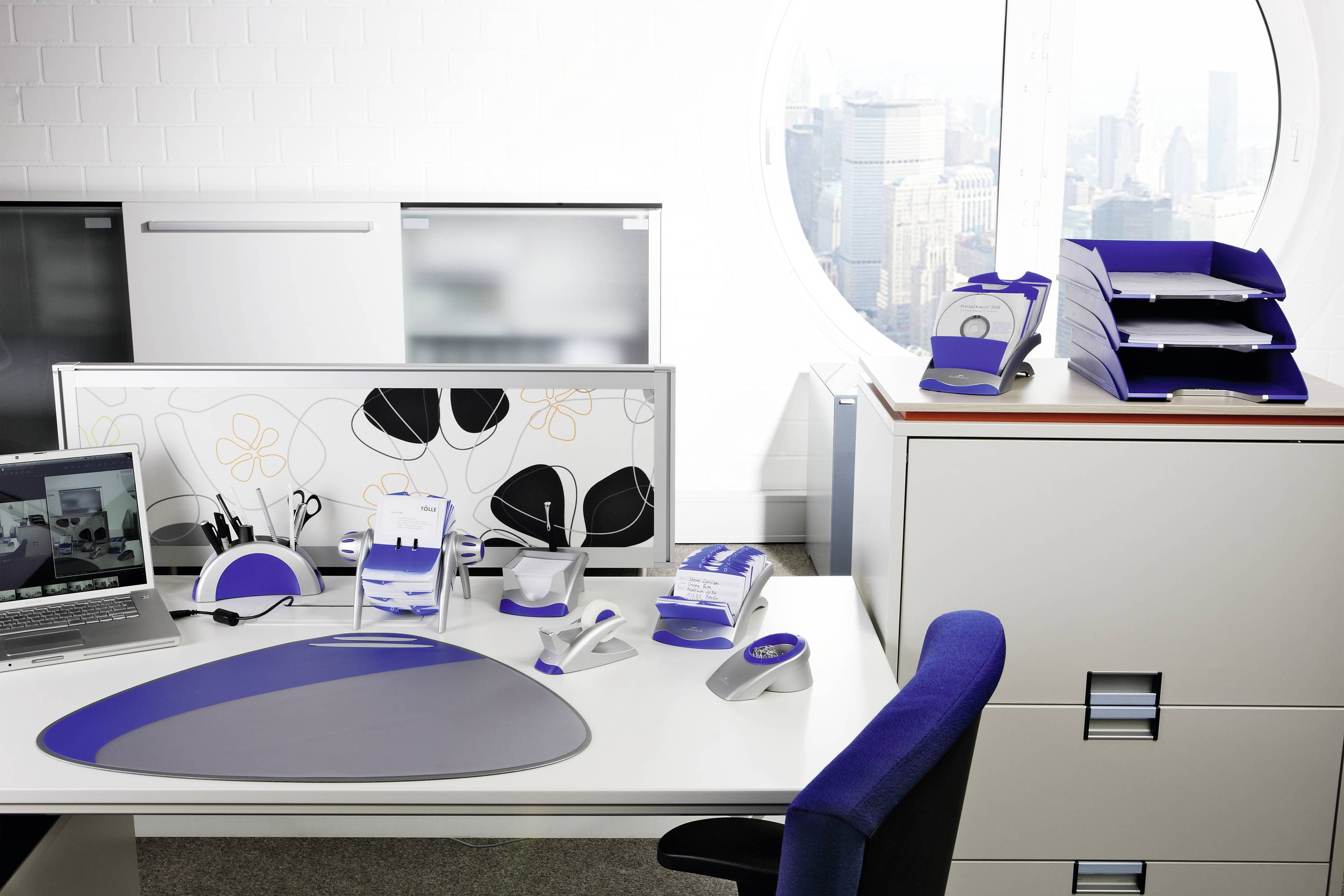 Ein ordentliches Büro mit Schreibtisch, Computer, Schreibwaren und Aktenablagen vor einem runden Fenster mit Stadtblick.