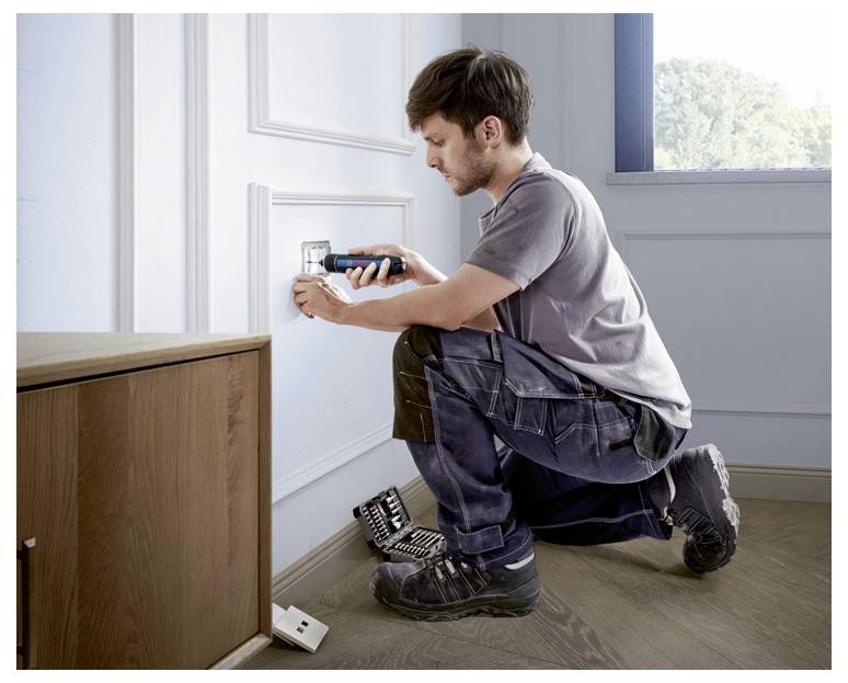 Ein Mann repariert eine Steckdose an einer Wand. Er kniet auf dem Boden, hält ein Werkzeug in der Hand, neben ihm liegen weitere Werkzeuge.