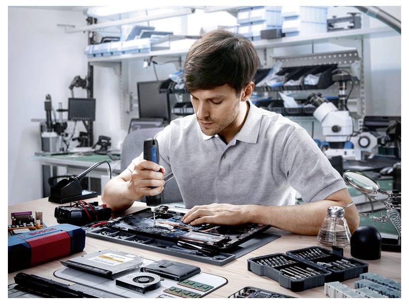 Ein Mann repariert ein elektronisches Gerät auf einem Tisch, umgeben von Werkzeugen und Ersatzteilen vor einem Laborhintergrund.