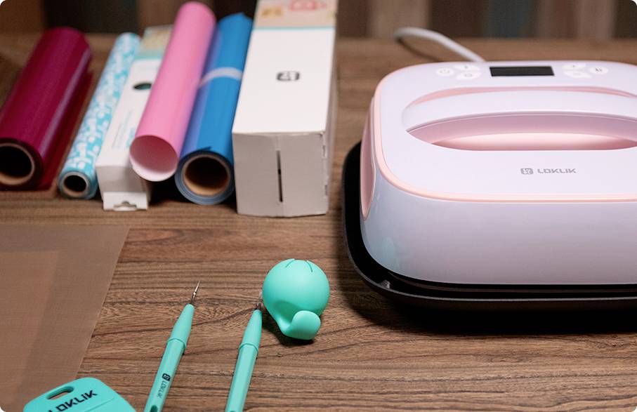 Ein rosa Heißpressgerät steht auf einem Tisch neben unterschiedlichen bunten Vinylrollen und Werkzeugen zur Bearbeitung von Materialien.