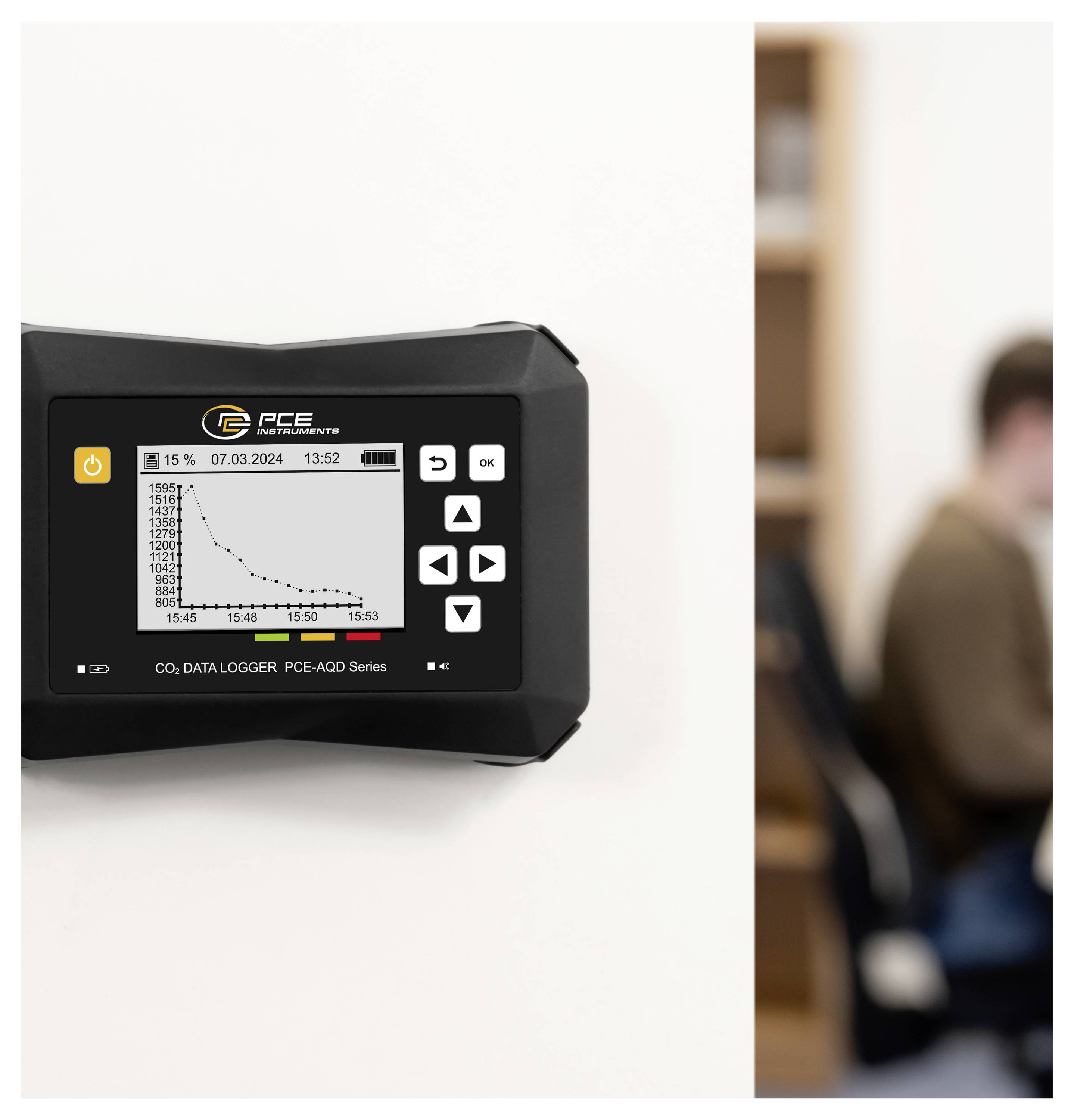An der Wand montiertes CO2-Datenerfassungsgerät, das ein Diagramm mit Abwärtstrend anzeigt, mit einer unscharfen Person im Hintergrund auf der rechten Seite.