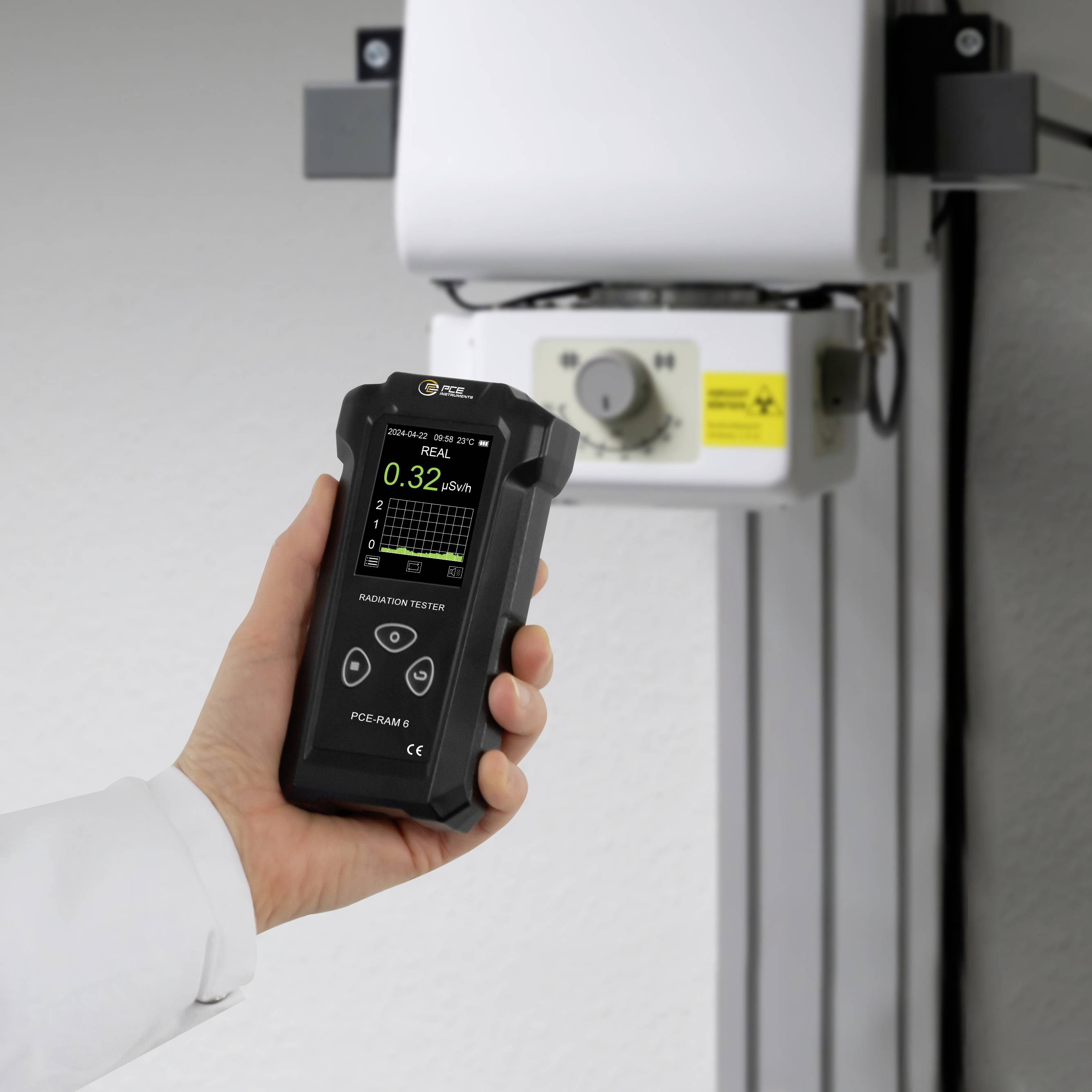 Eine Hand hält ein Strahlungsmessgerät, das 0,32 µSv/h anzeigt, vor einem Röntgengerät.