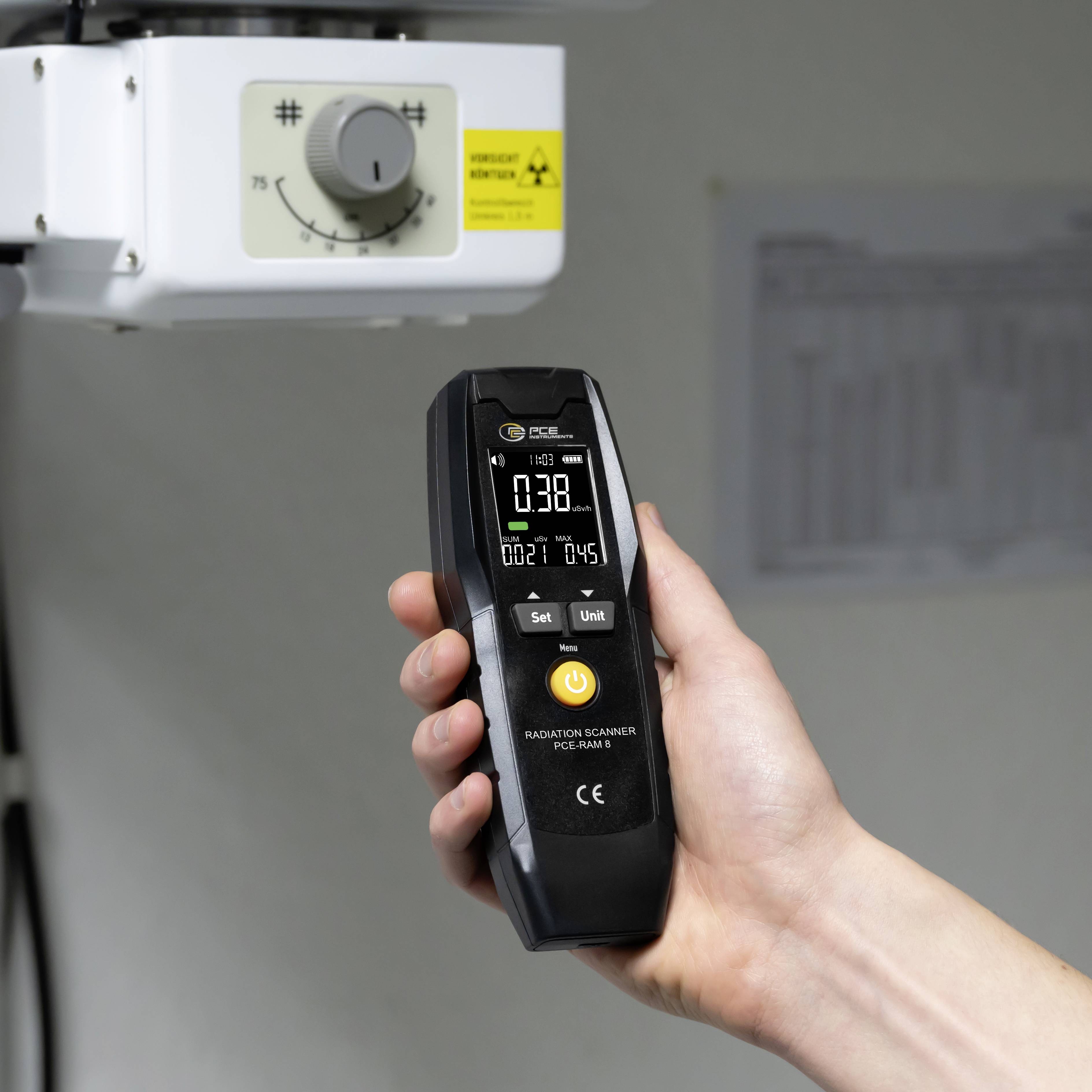 Eine Hand hält einen Geigerzähler, der unter einem Röntgengerät eine Strahlungsmessung von 0,38 µSv/h anzeigt.