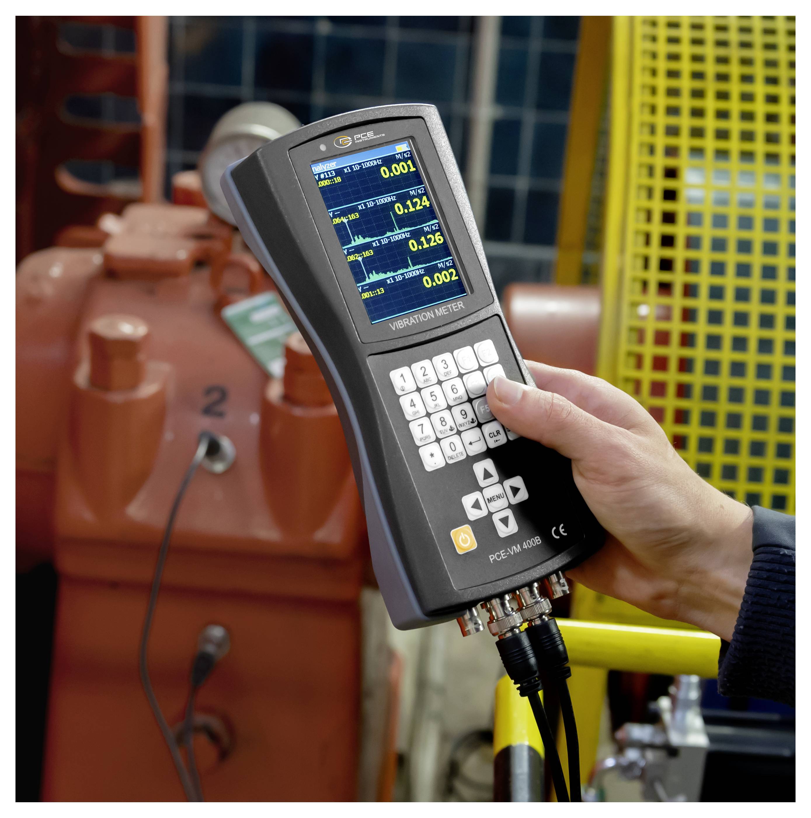 Hand hält ein Vibrationsmessgerät mit Live-Messwerten, Messequipment in einer industriellen Umgebung in der Nähe einer Maschine oder eines Motors.