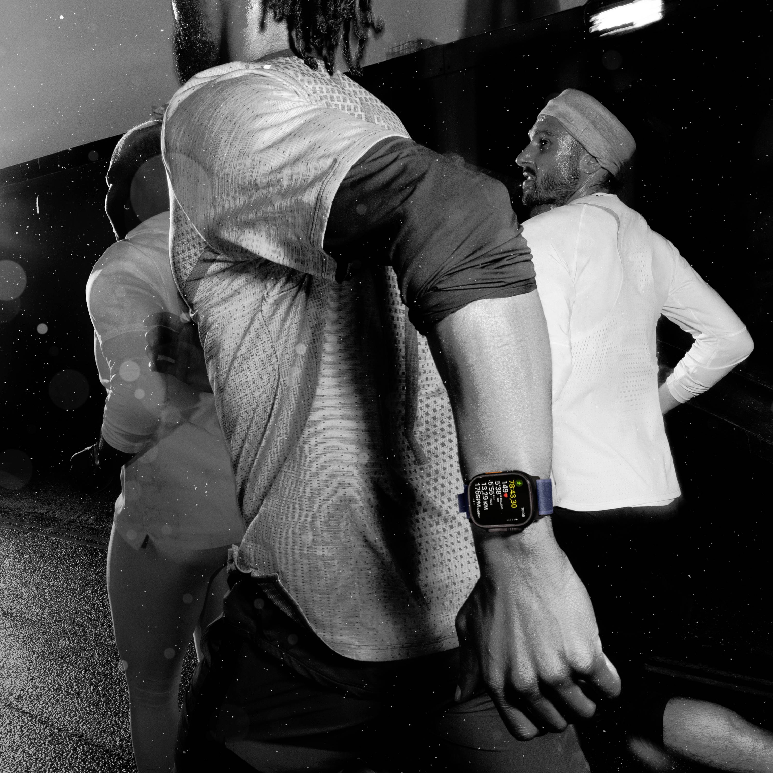 Eine Person mit einer Smartwatch joggt bei Nacht. Die Uhr zeigt Gesundheitsdaten an. Personen im Hintergrund laufen ebenfalls.