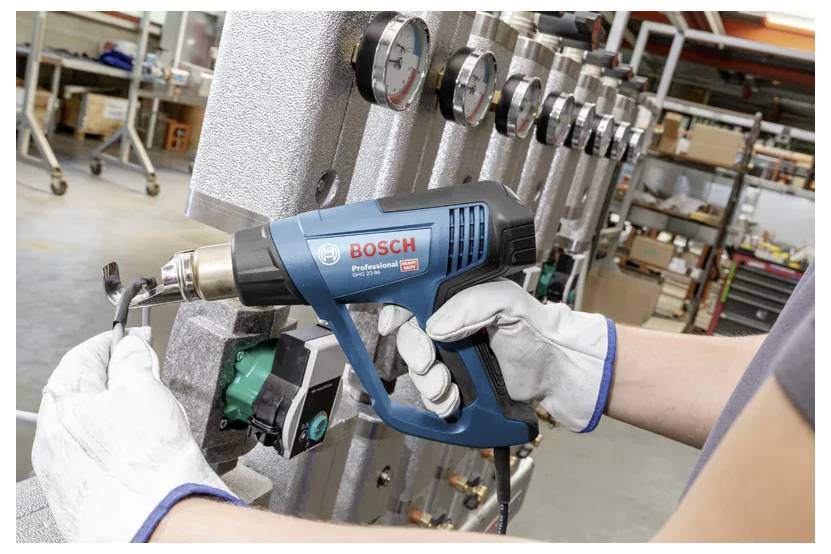Eine Person mit Handschuhen bedient einen Bosch-Heißluftfön an einer Rohrisolierung, mit industriellen Messgeräten im Hintergrund, in einer Werkstattumgebung.