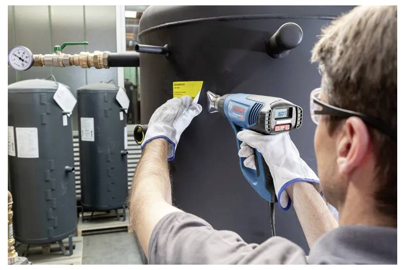 Eine Person mit Handschuhen verwendet einen Heißluftföhn, um ein gelbes Etikett auf einen großen Industrietank anzubringen, wobei weitere Tanks im Hintergrund zu sehen sind.
