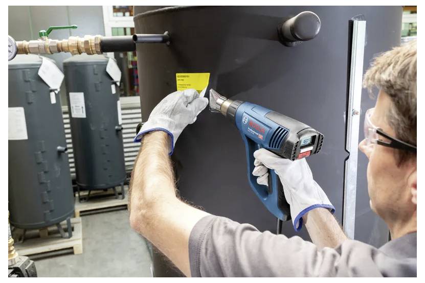 Eine Person, die einen Heißluftföhn auf ein gelbes Etikett an einem großen zylindrischen Tank richtet und dabei Handschuhe und Schutzbrille in einer industriellen Umgebung trägt.