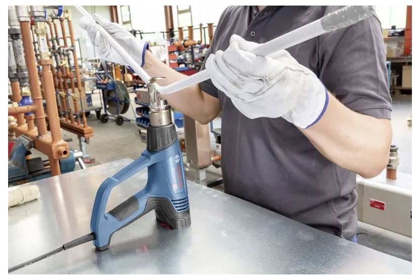 Eine Person mit Handschuhen verwendet einen blauen Heißluftfön, um ein graues Kunststoffrohr auf einem Werkstatttisch zu biegen, während verschiedene Werkzeuge im Hintergrund zu sehen sind.