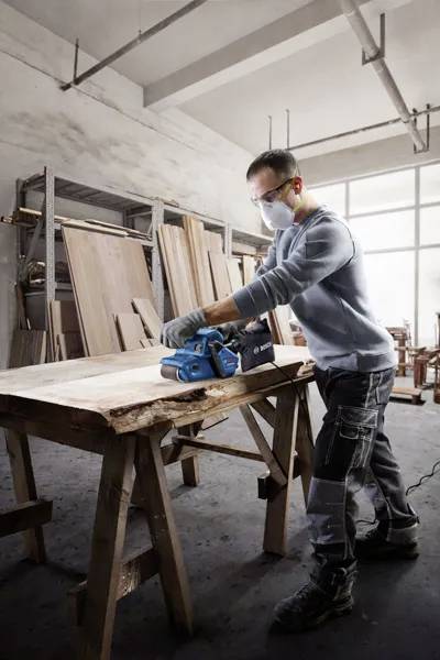 Eine Person mit Sicherheitsausrüstung bedient einen Exzenterschleifer an einem Holzbrett in einer Werkstatt. Im Hintergrund sind verschiedene Holzplatten zu sehen.