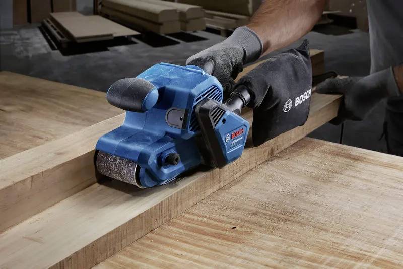 Eine Person verwendet einen blauen Bosch-Bandschleifer auf einem Holzbrett mit Präzision und veranschaulicht damit Holzbearbeitungswerkzeuge und Handwerkskunst in Aktion.