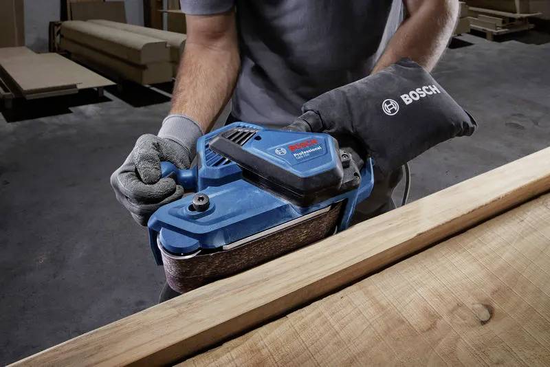 Eine Person verwendet einen blauen Bosch-Bandschleifer auf einem Holzbrett in einer Werkstatt.