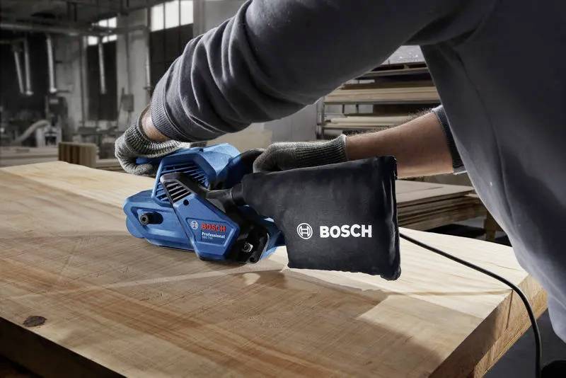 Eine Person, die einen blauen Bosch-Bandschleifer auf einer Holzoberfläche in einer Werkstattumgebung verwendet und dabei die Leistungsfähigkeit und Vielseitigkeit des Werkzeugs hervorhebt.