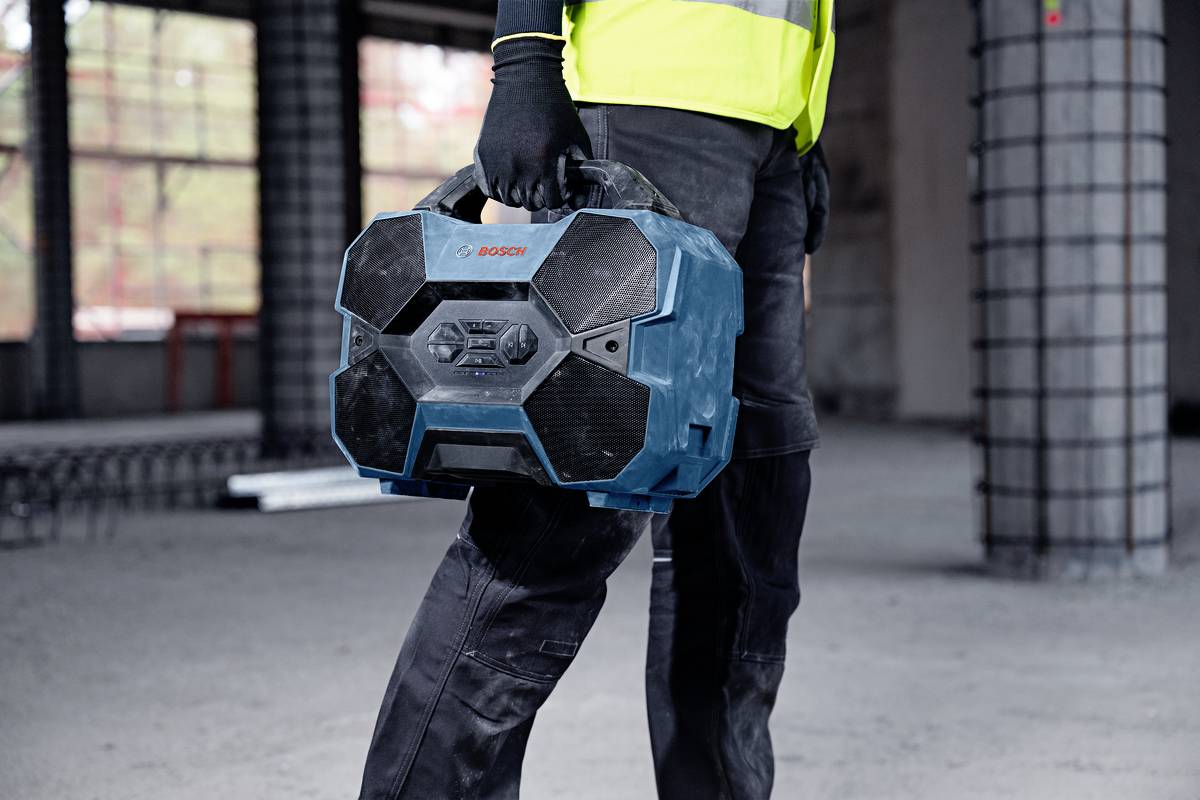 Ein Bauarbeiter trägt eine blaue, sechseckige Werkzeugkiste in einem unfertigen Gebäude. Der Arbeiter trägt eine reflektierende Weste.