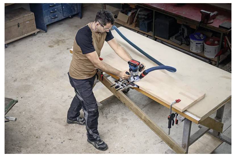 Eine Person arbeitet mit einem Elektrowerkzeug an einem großen Holzstück in einer Werkstatt. Im Hintergrund sind Werkbänke und Werkzeuge zu sehen.
