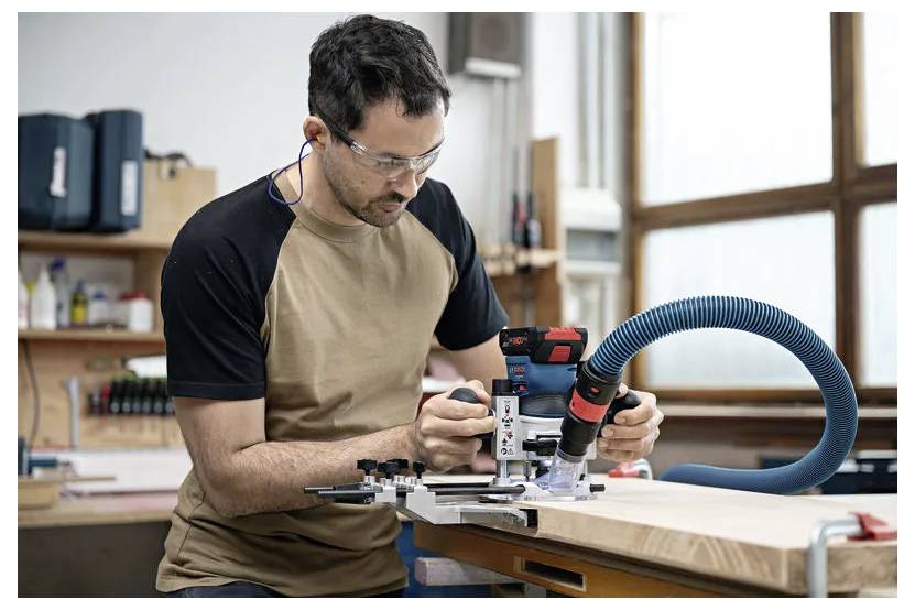 Ein Mann mit Schutzbrille und Gehörschutzstöpseln bedient ein Elektrowerkzeug an einer Holzwerkbank in einer Werkstatt.