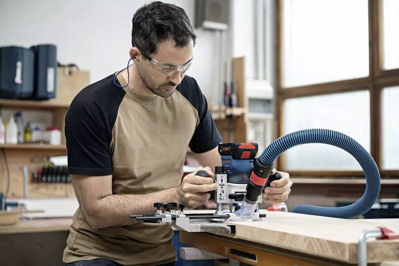 Ein Mann mit Schutzbrille bearbeitet ein Holzstück mit einem Elektrowerkzeug in einer Werkstatt. Im Hintergrund sind Regale zu sehen.
