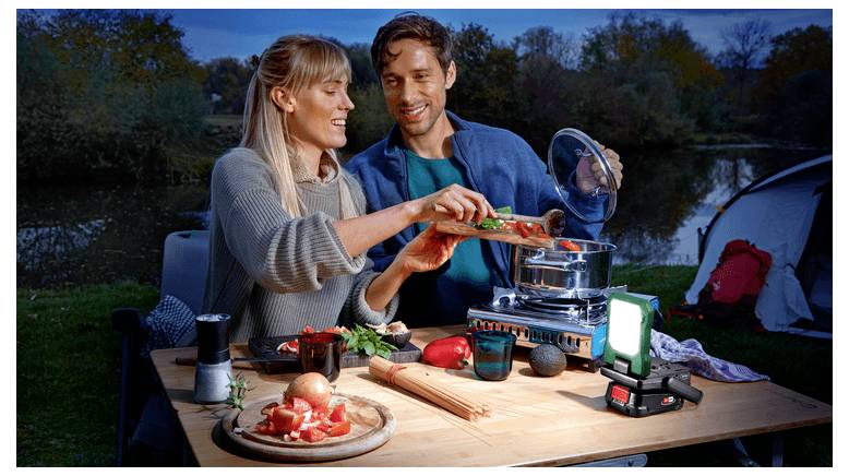 Paar kocht im Freien auf einem Campingplatz und gibt Lebensmittel in einen Topf auf einem tragbaren Kocher neben einer brennenden Laterne und Campingausrüstung.