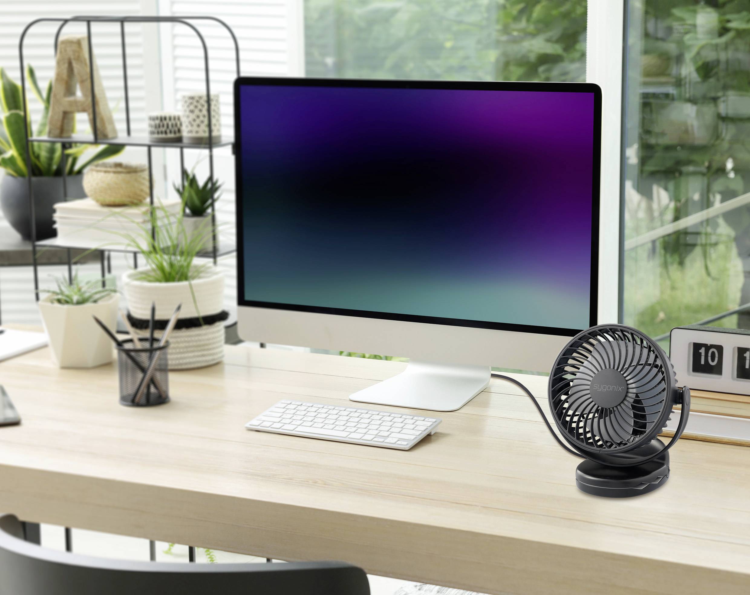 Ein moderner Schreibtisch mit einem großen Computermonitor, kabelloser Tastatur und Maus. Rechts steht ein kleiner Ventilator, umgeben von Pflanzen.