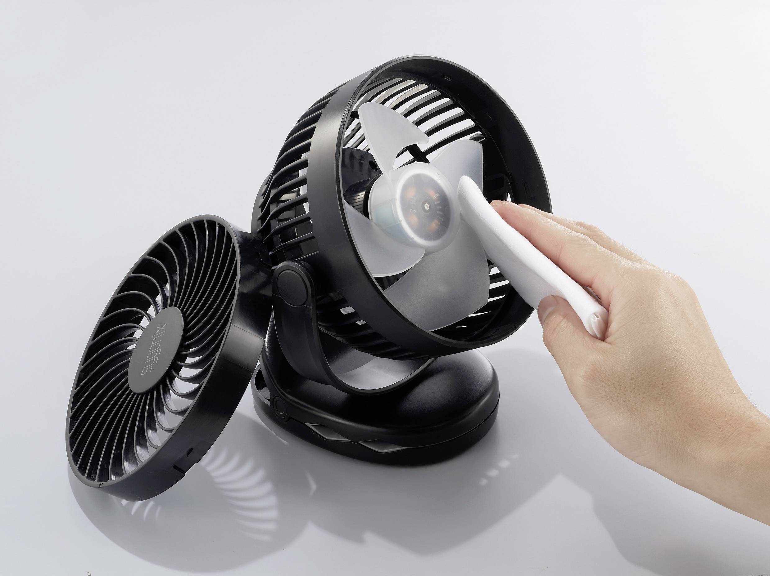 Eine Hand reinigt die Rotorblätter eines kleinen, schwarz-grauen Ventilators mit einer weißen Bürste. Im Hintergrund ist ein zweiter Ventilator.