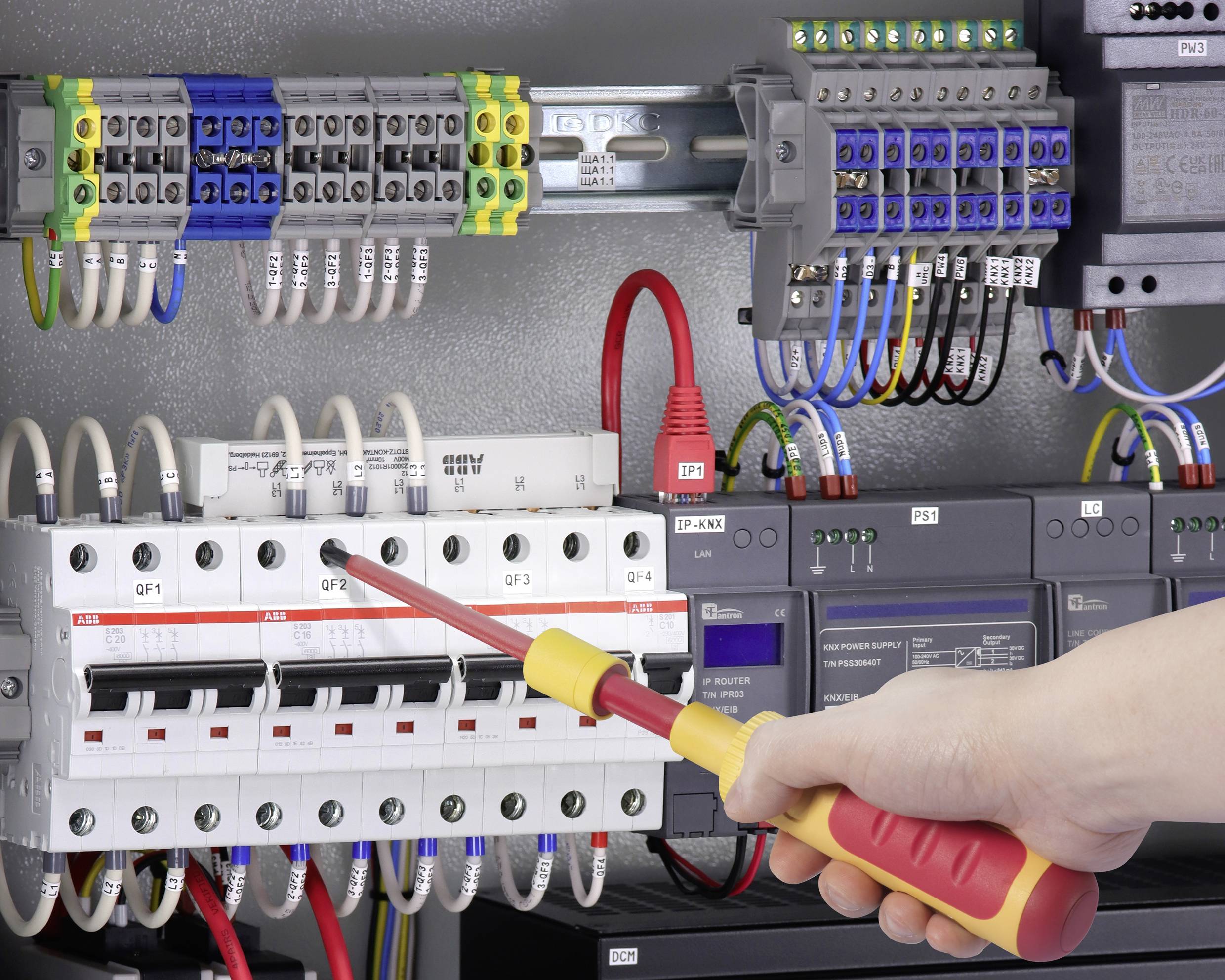 Eine Person arbeitet mit einem Schraubenzieher an einer Schalttafel mit vielen elektrischen Schaltungen und farbigen Kabeln.