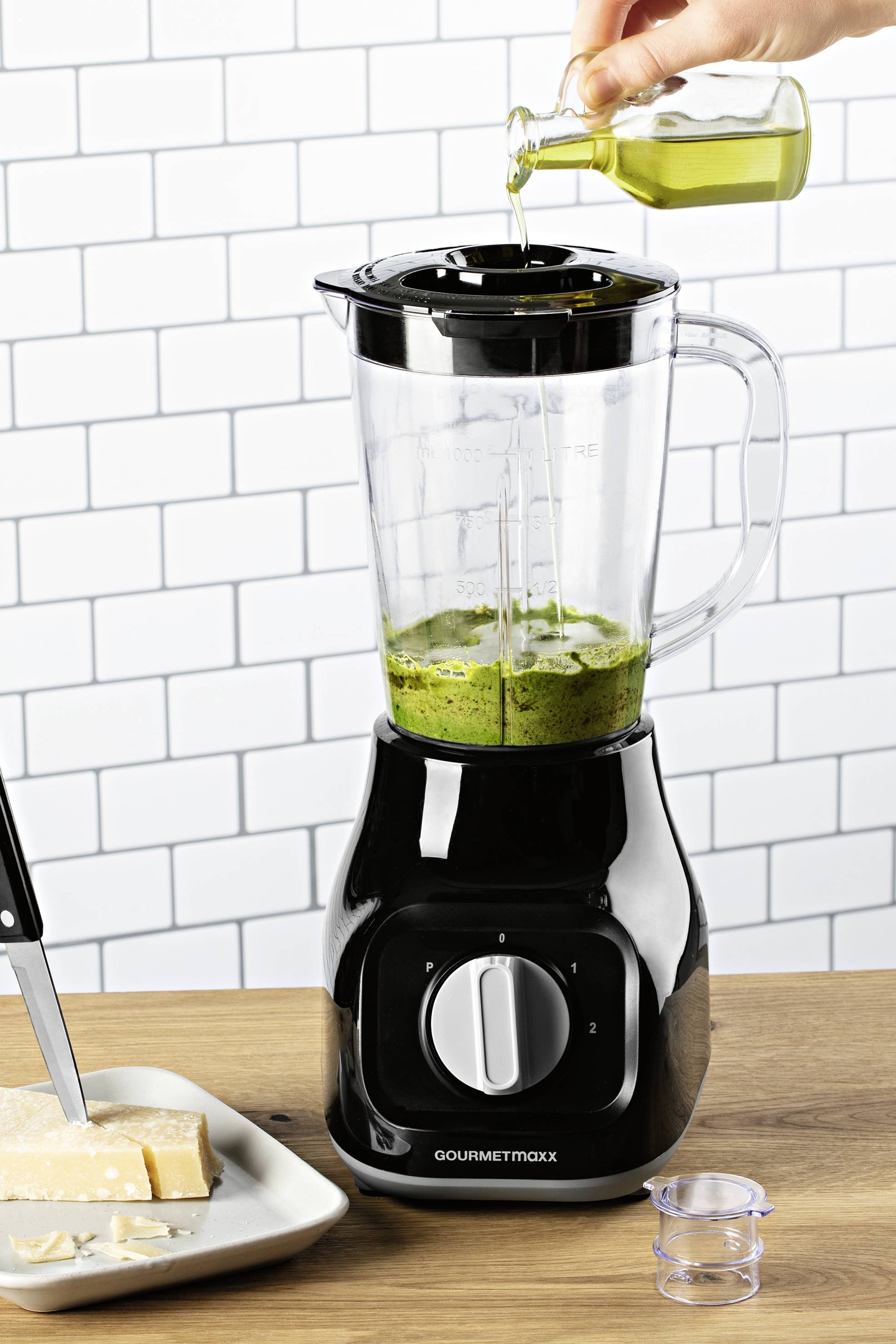 Eine Person gießt Öl in einen Mixer mit einer grünen Mischung. Daneben ein Teller mit Käsewürfeln und einem Messer auf einem Holztisch.