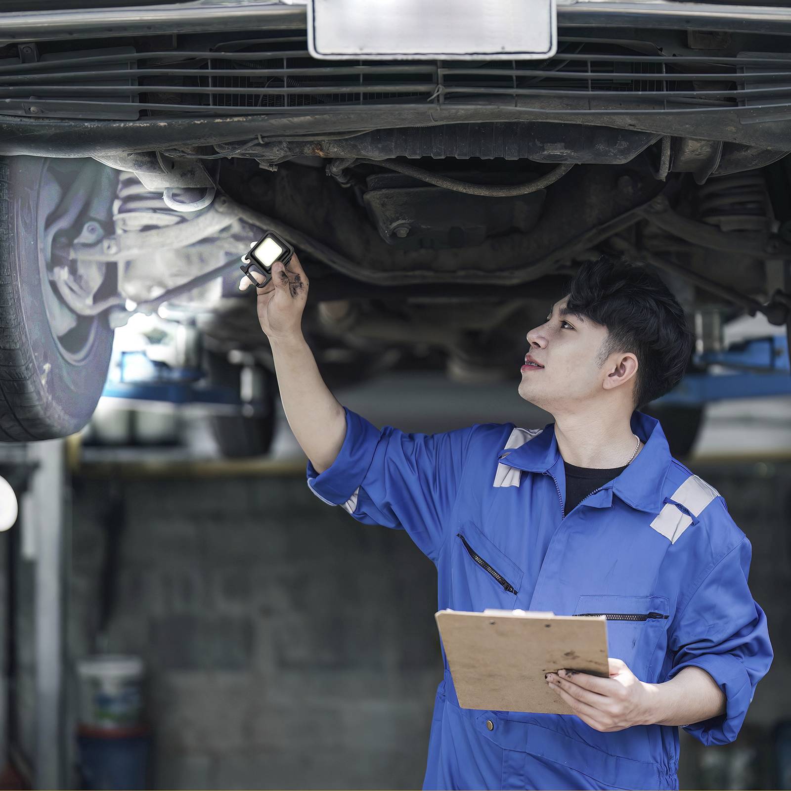Mechaniker in blauer Arbeitskleidung inspiziert ein Auto von unten in einer Werkstatt, während er Notizen auf einem Klemmbrett macht.