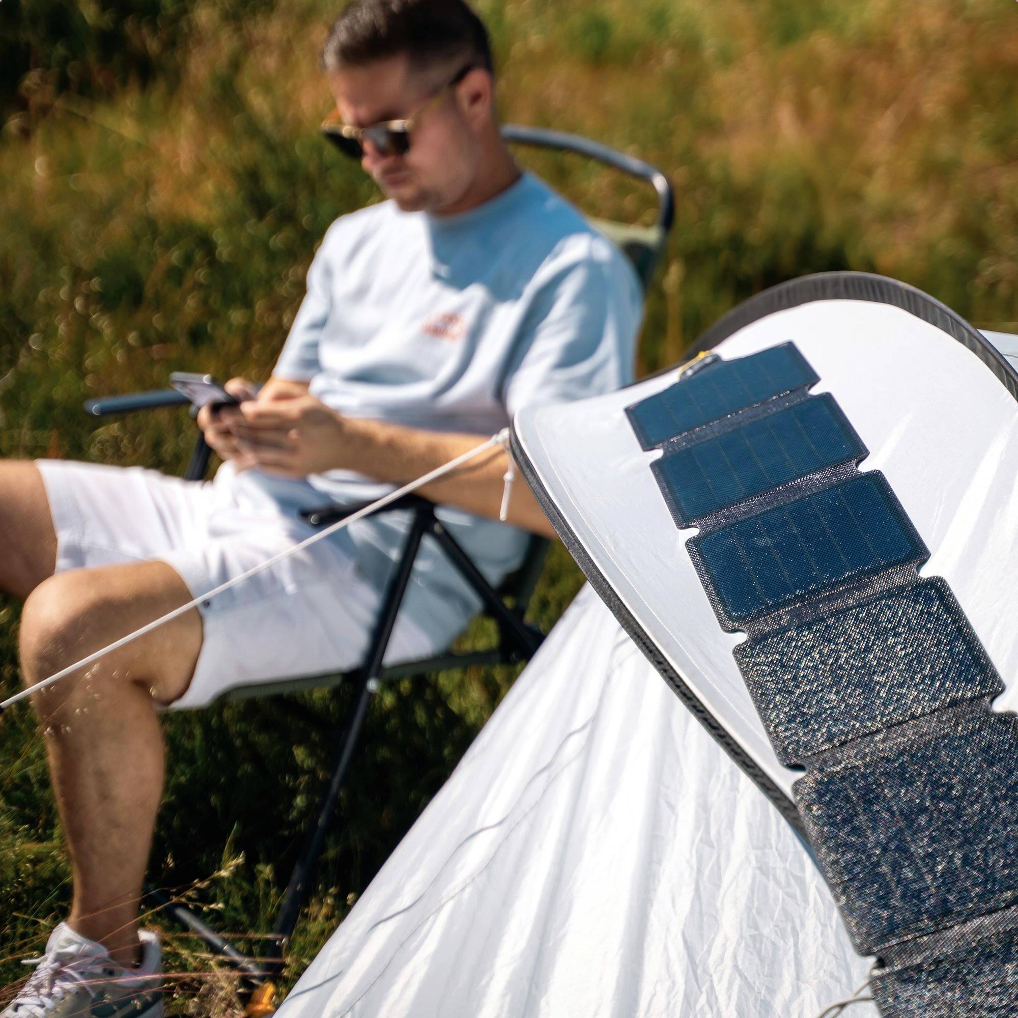 Ein Mann sitzt im Freien und benutzt ein Smartphone, während er neben einer Solarpanel-bedeckten Zeltplane entspannt.