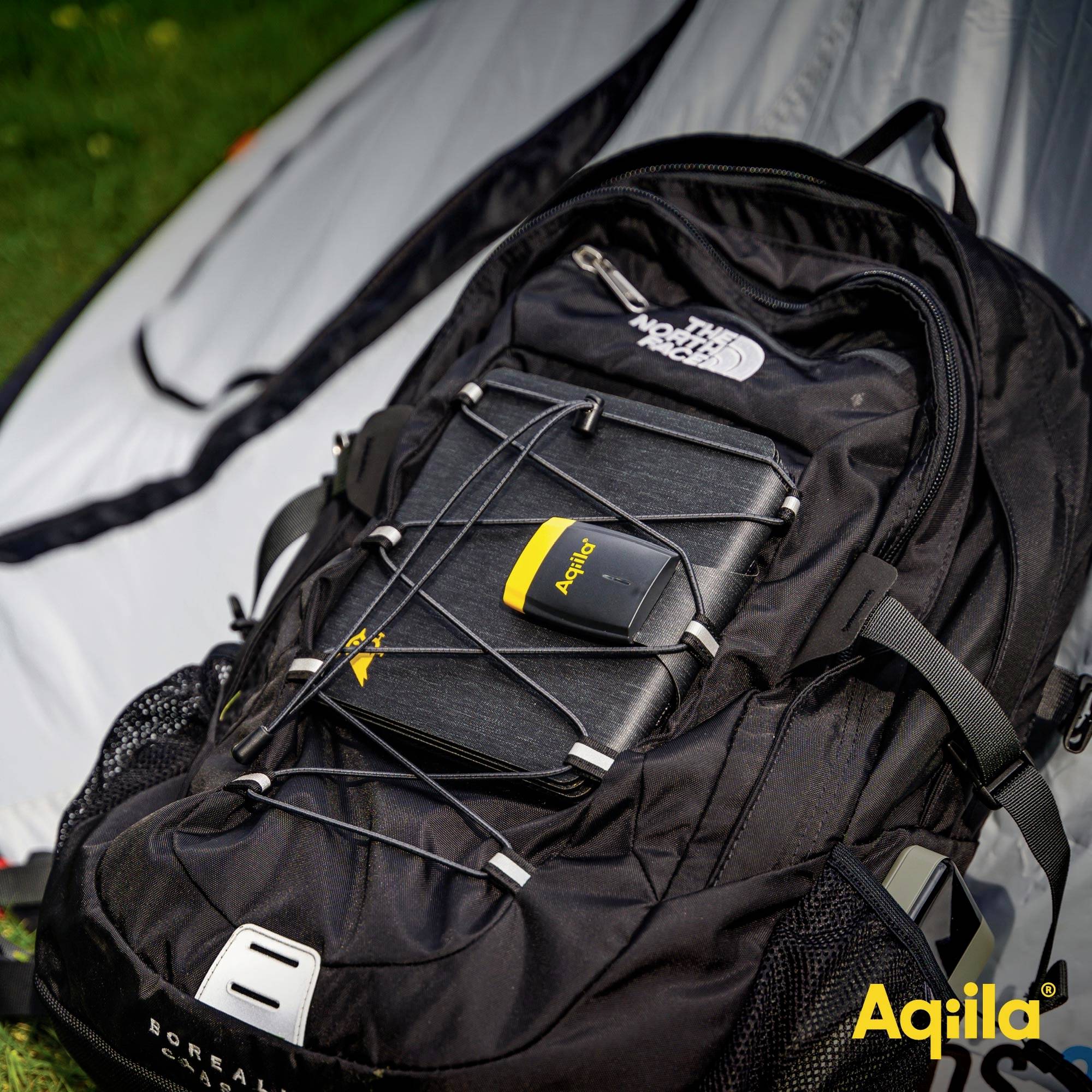 Ein Rucksack liegt vor einem Zelt. Ein elektronisches Gerät ist oben am Rucksack befestigt. Markenlogo 'Aquila' unten rechts.