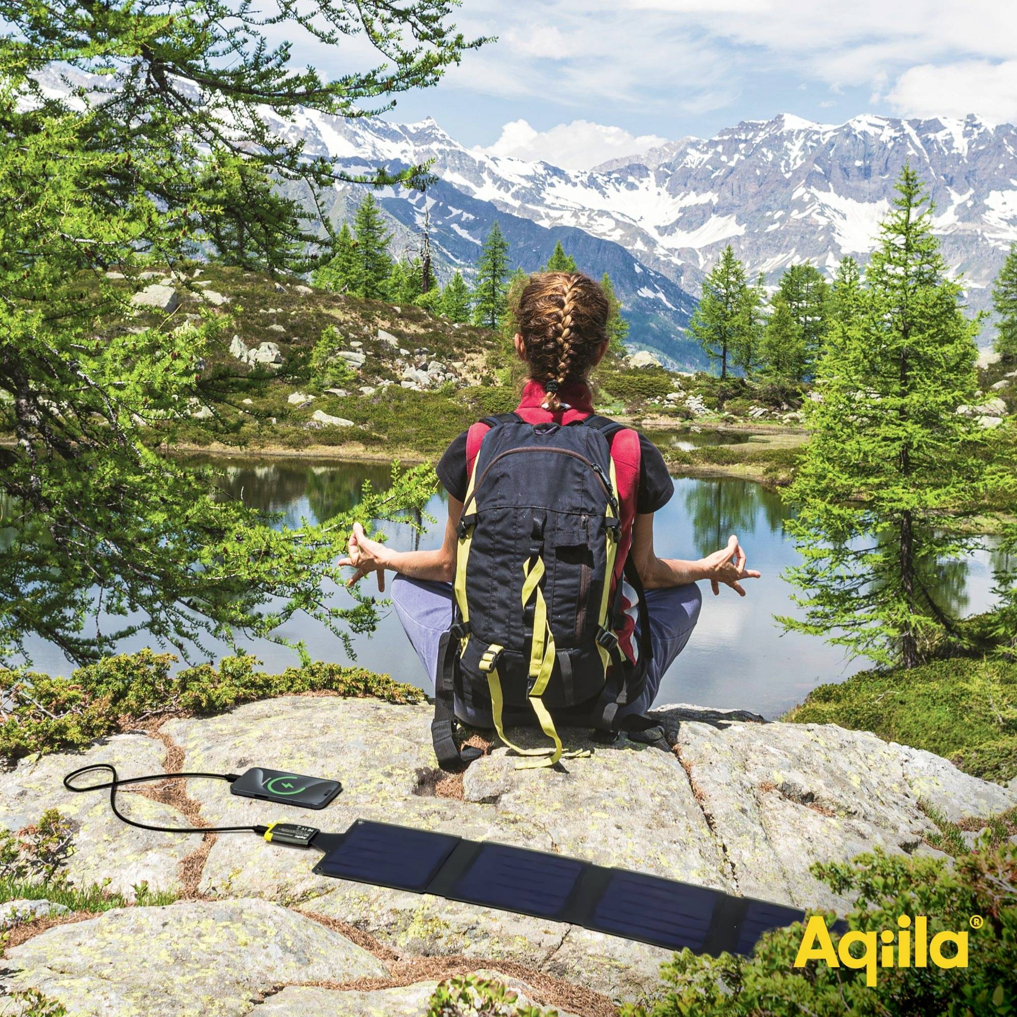 Eine Person sitzt in Meditationshaltung auf einem Felsen in den Bergen. Ein Solarladegerät lädt ein Smartphone auf. Im Hintergrund Berge und Bäume.
