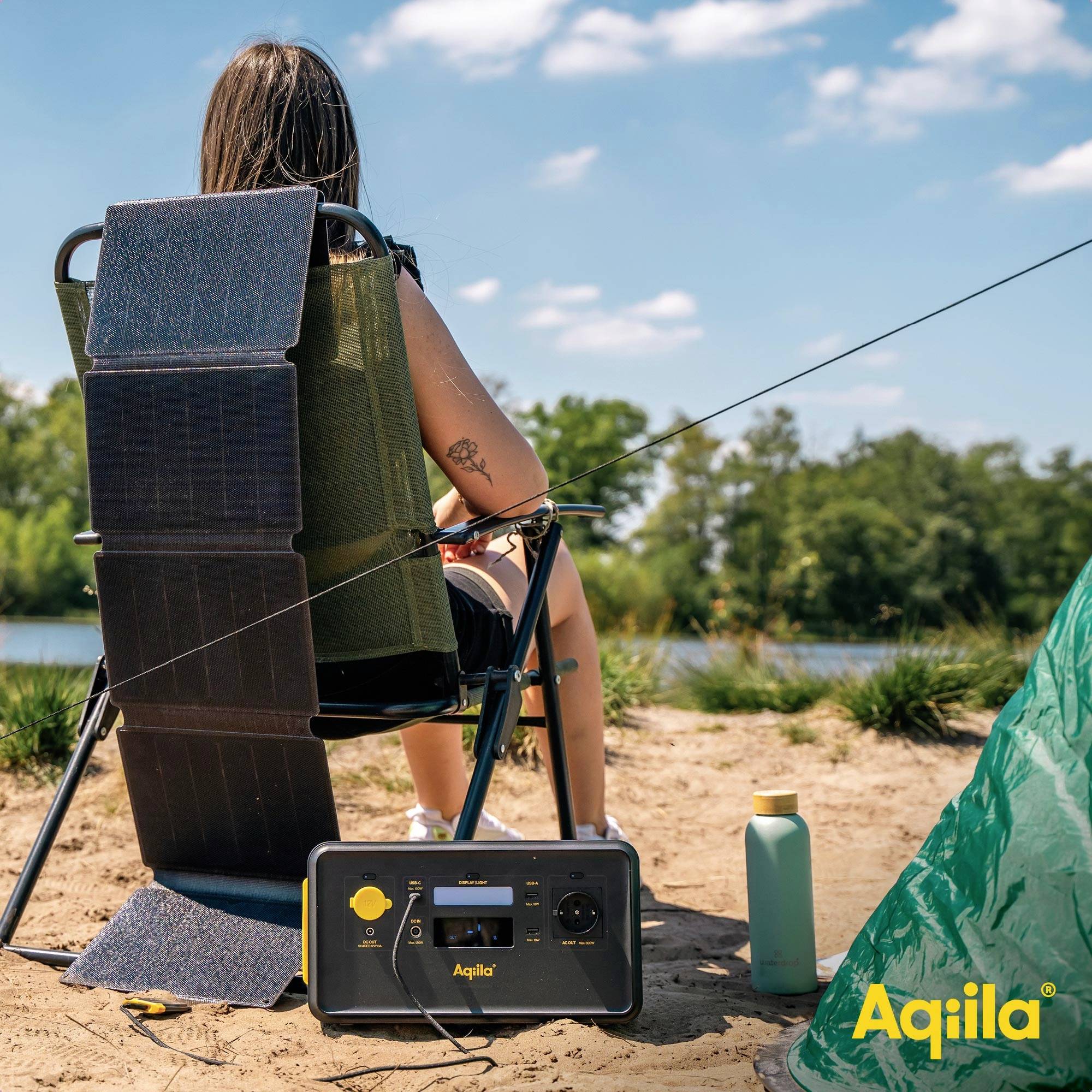 Eine Person sitzt am See, lädt ein Gerät mit einem tragbaren Solarpanel auf. Im Vordergrund ist eine Wasserflasche.