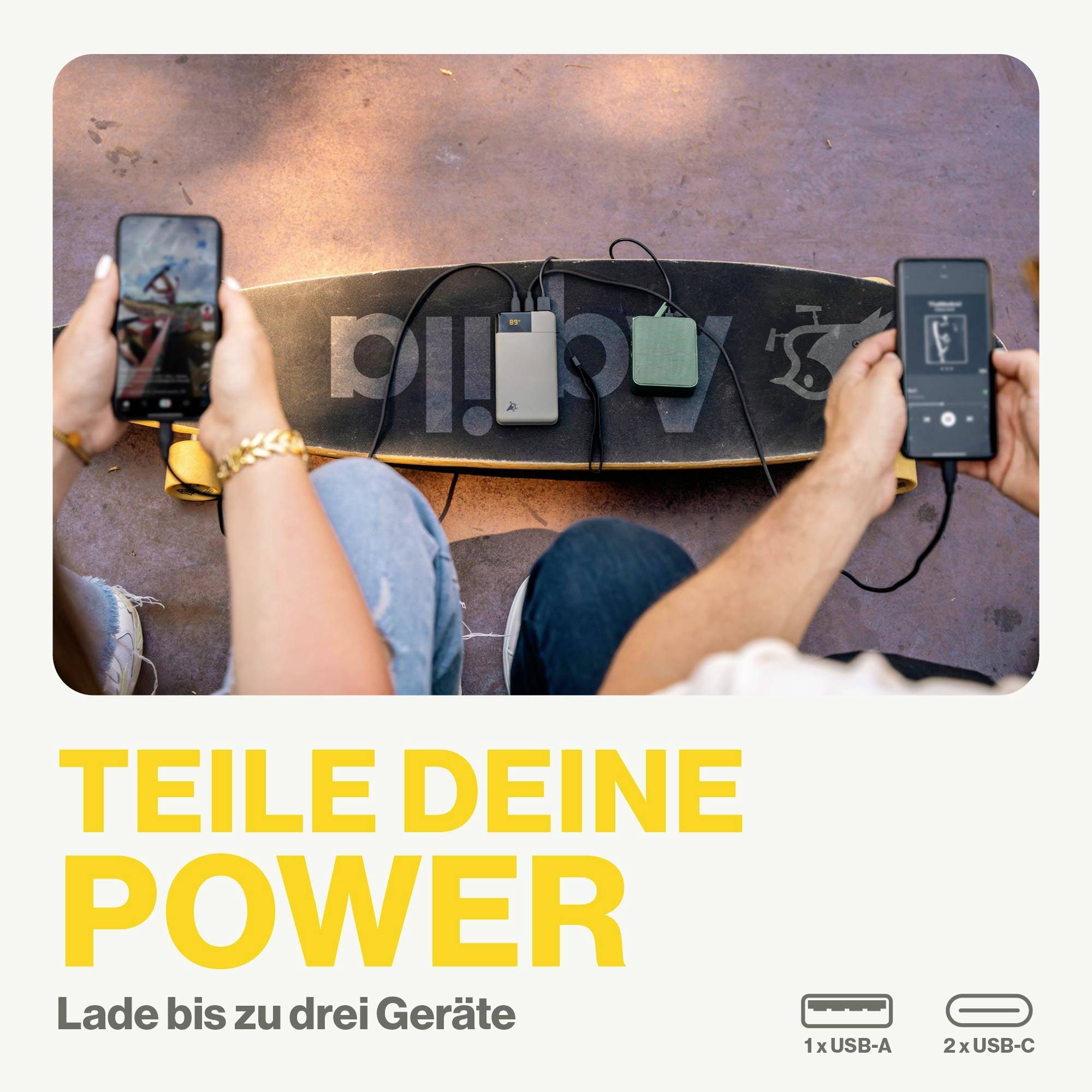Zwei Personen laden ihre Geräte mit Powerbanks auf, die auf einem Skateboard liegen. Text: 'Teile deine Power. Lade bis zu drei Geräte'.