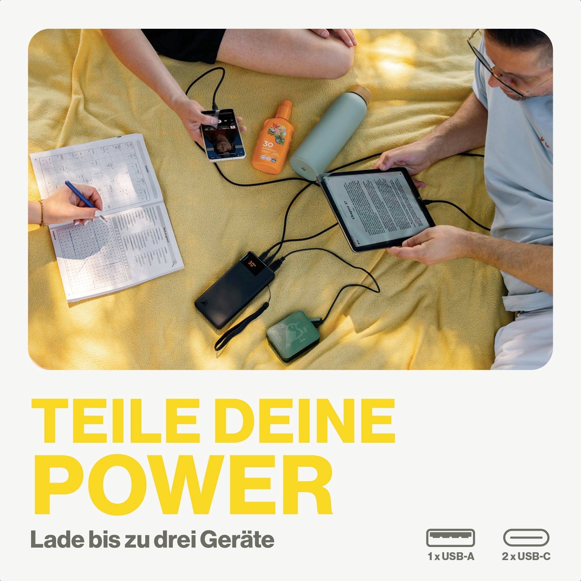 Drei Personen sitzen auf einer gelben Decke im Freien und laden ein Handy, ein Tablet und eine Musikbox mit einem Solarpanel auf. Text: 'TEILE DEINE POWER'.
