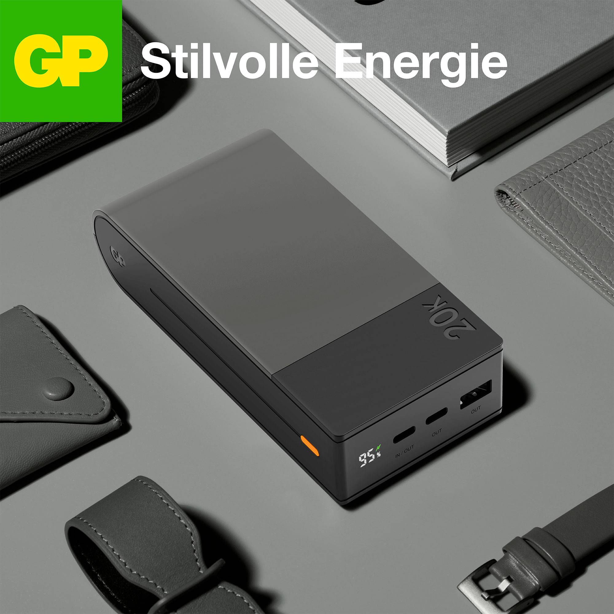Eine Powerbank mit digitaler Anzeige und zwei USB-Anschlüssen liegt stilvoll auf einem Tisch neben einer Ledertasche und einem Notizbuch.