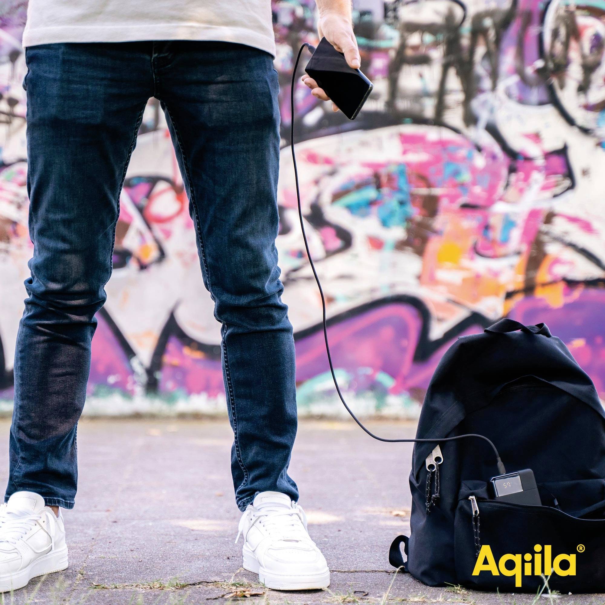 Eine Person hält ein Handy und steht vor einer bunten Graffiti-Wand. Ein Rucksack liegt am Boden, verbunden mit einem Ladekabel.