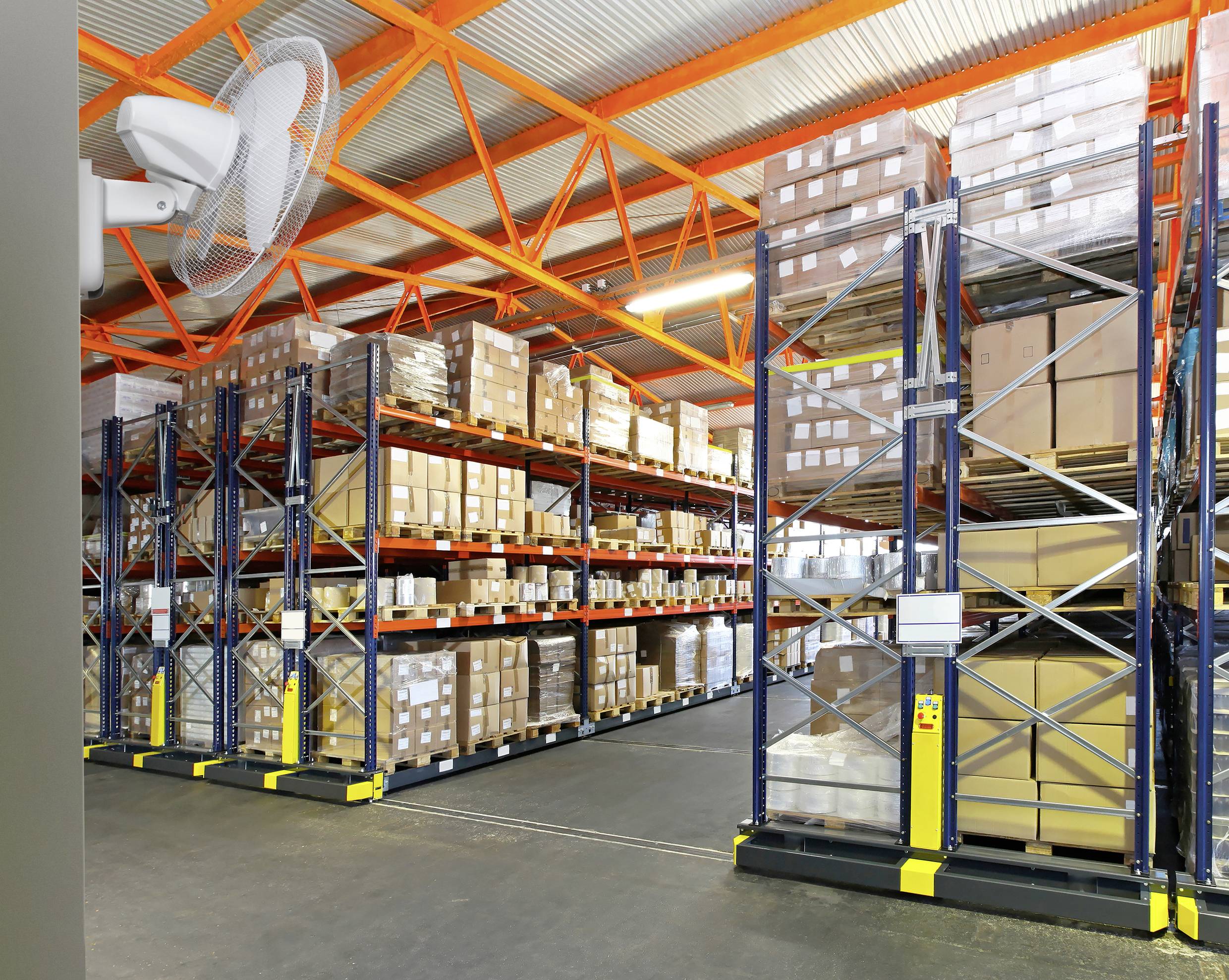 Ein Lagerhaus mit hohen Regalen voller Kartons und Pakete. Ein Ventilator an der Wand links sorgt für Belüftung.