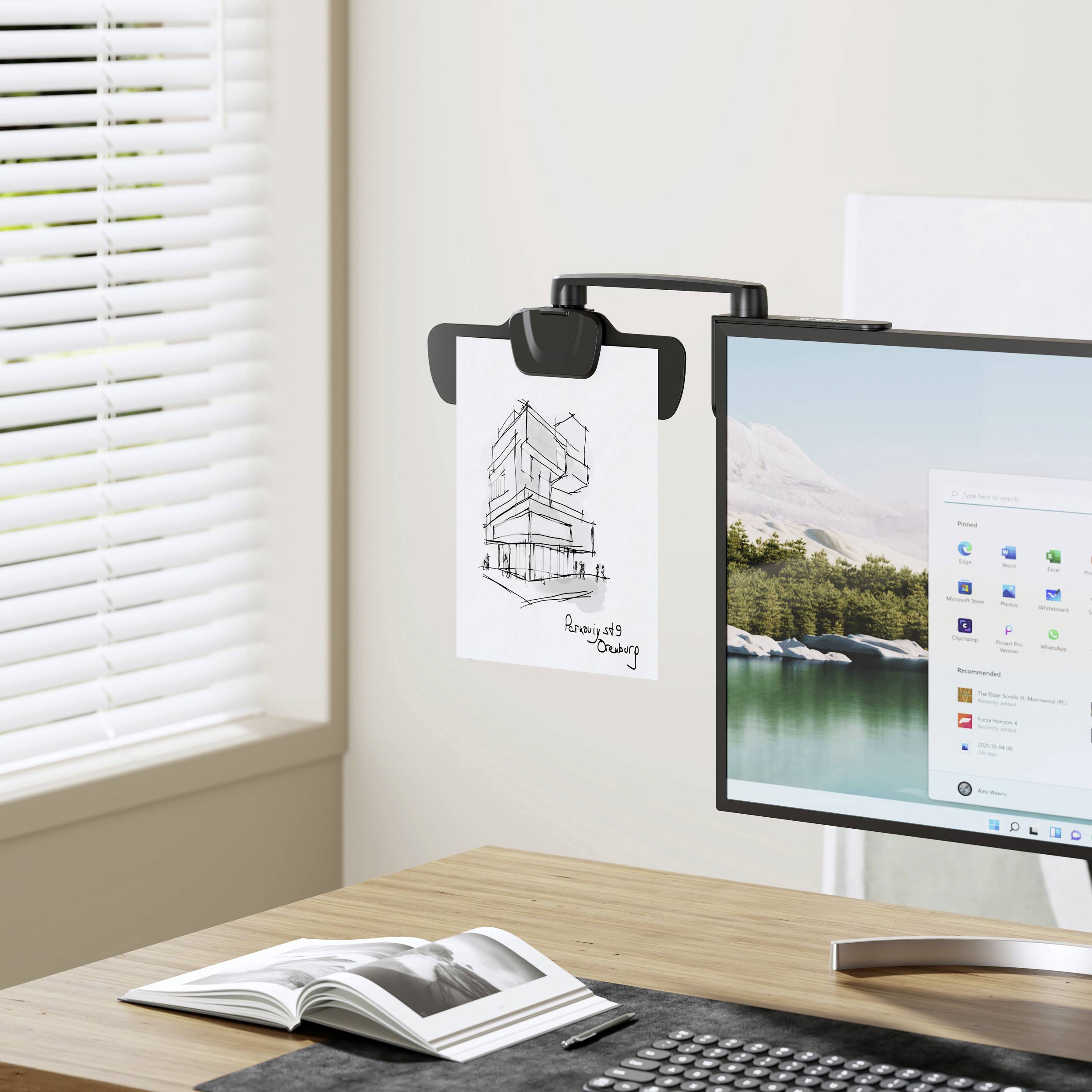 Ein Computermonitor zeigt die Windows-Benutzeroberfläche. Daneben hängt eine Zeichnung eines modernen Gebäudes am Monitor. Im Hintergrund ist ein Fenster.