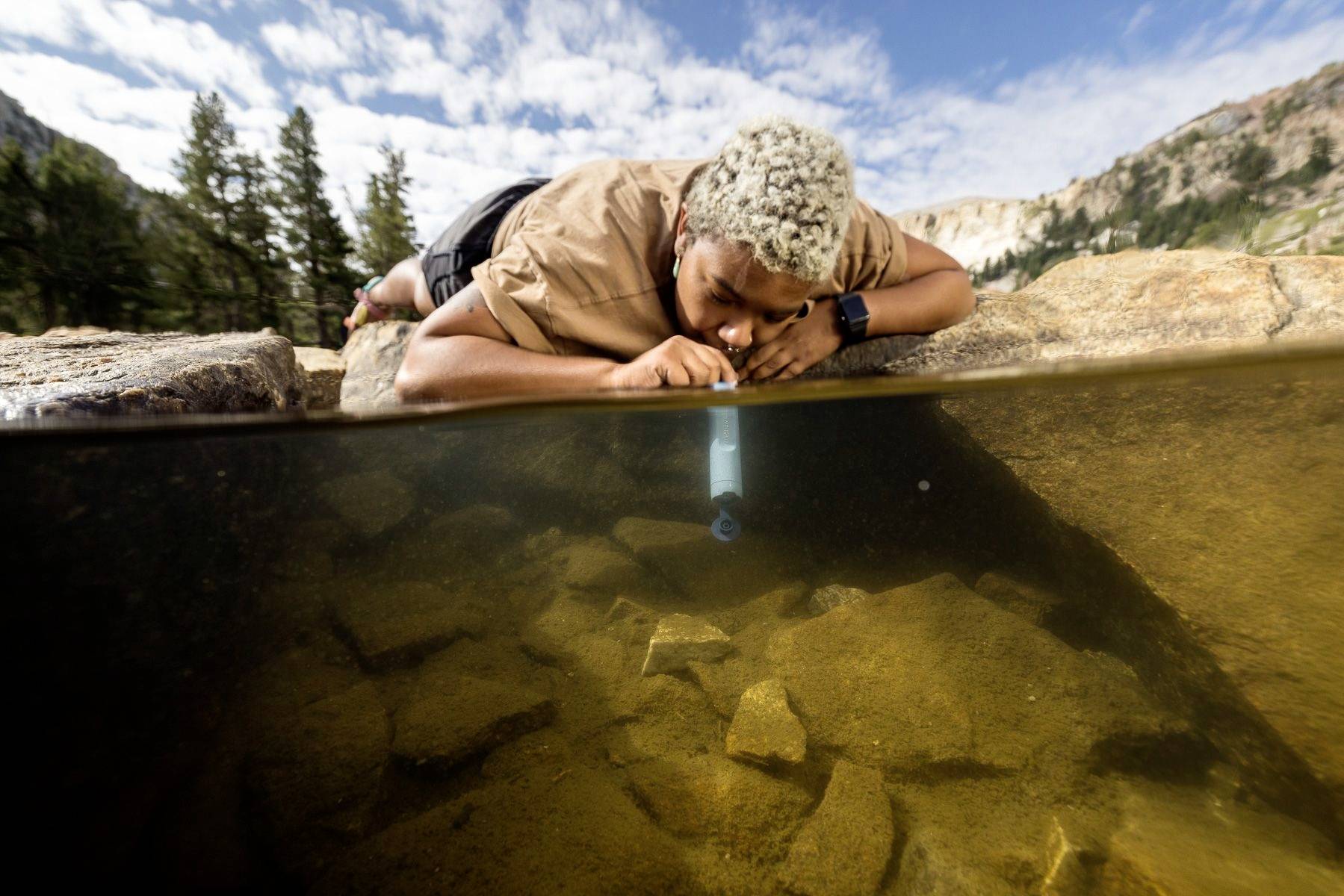 Eine Person trinkt Wasser aus einem Bach in einer Berglandschaft mit Hilfe eines Wasserfilters.