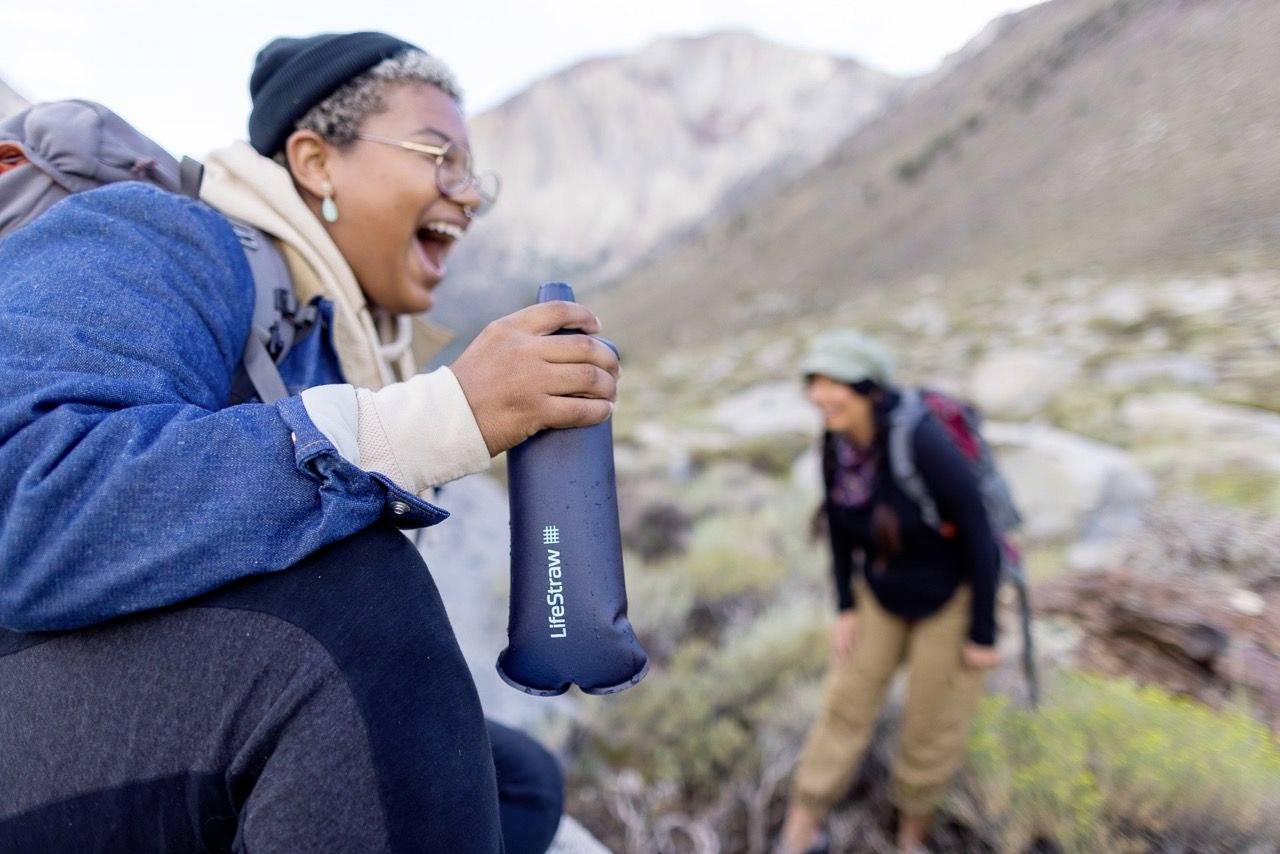 Eine Person lacht und hält eine Wasserflasche, während sie mit einer anderen Person im Hintergrund in einer bergigen Landschaft wandert.