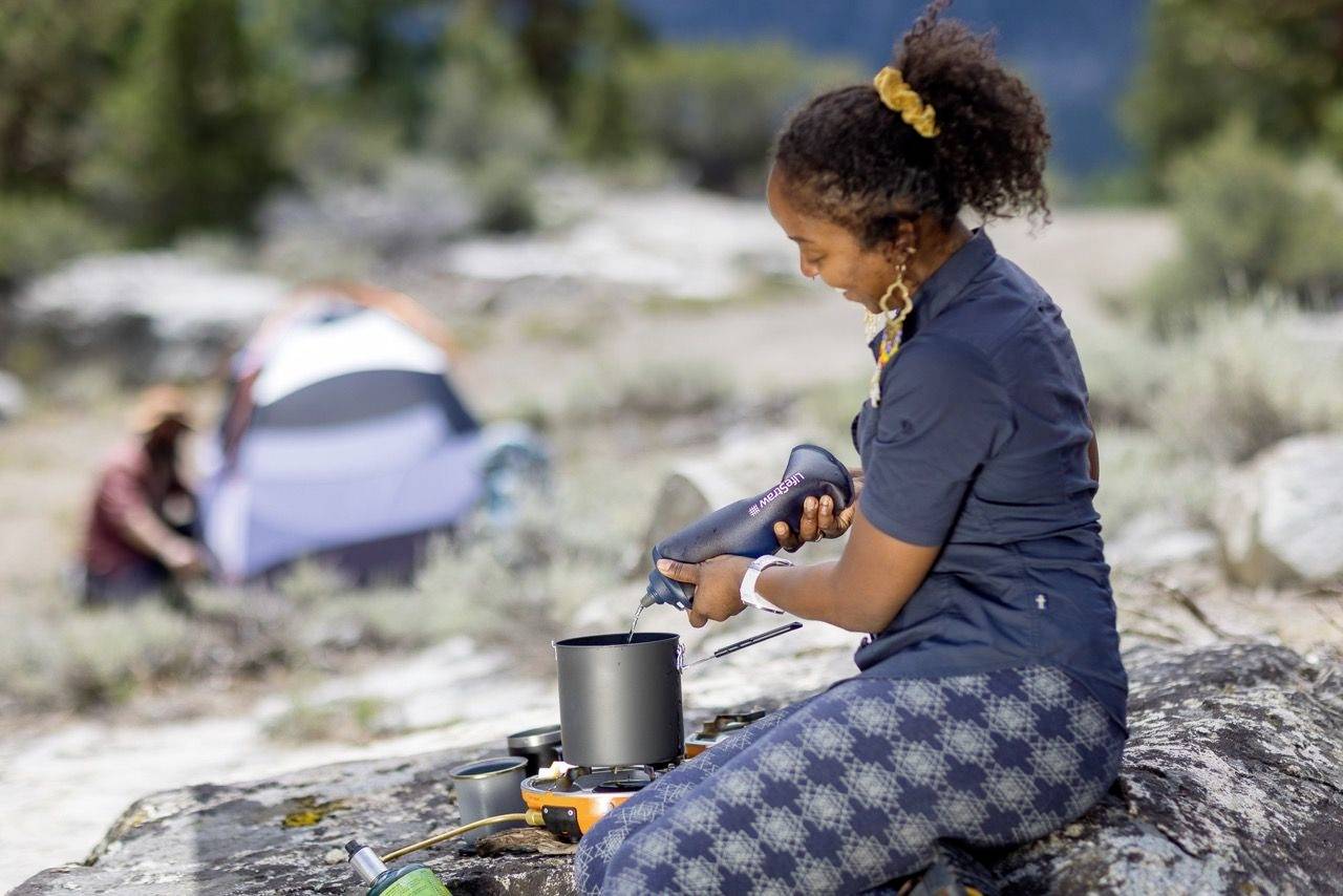 Eine Person kocht mit einem tragbaren Herd im Freien, im Hintergrund ein Zelt. Es ist Tag und die Szene vermittelt Campingaktivitäten.