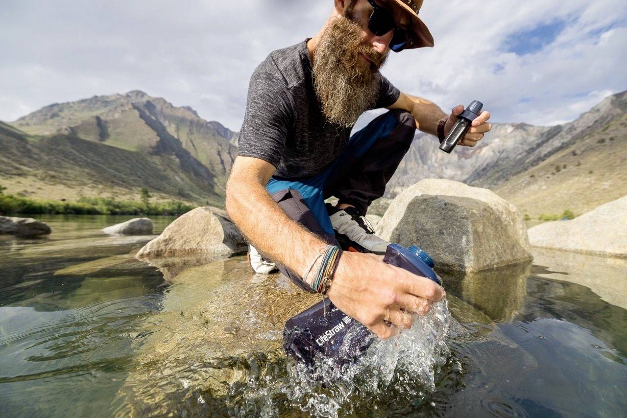 Mann mit langem Bart kniet auf einem Felsen am Fluss und füllt eine Wasserflasche. Er trägt Outdoor-Kleidung, Hut und Sonnenbrille. Berglandschaft im Hintergrund.