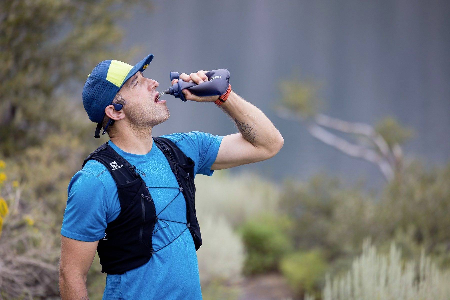 Ein Mann in Sportkleidung trinkt aus einer Wasserflasche, während er draußen in einer natürlichen Umgebung steht.