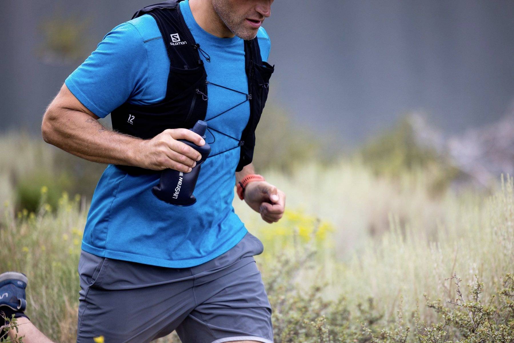 Person in blauem Sporthemd und grauer Hose läuft mit einer Wasserflasche durch eine natürliche Landschaft.