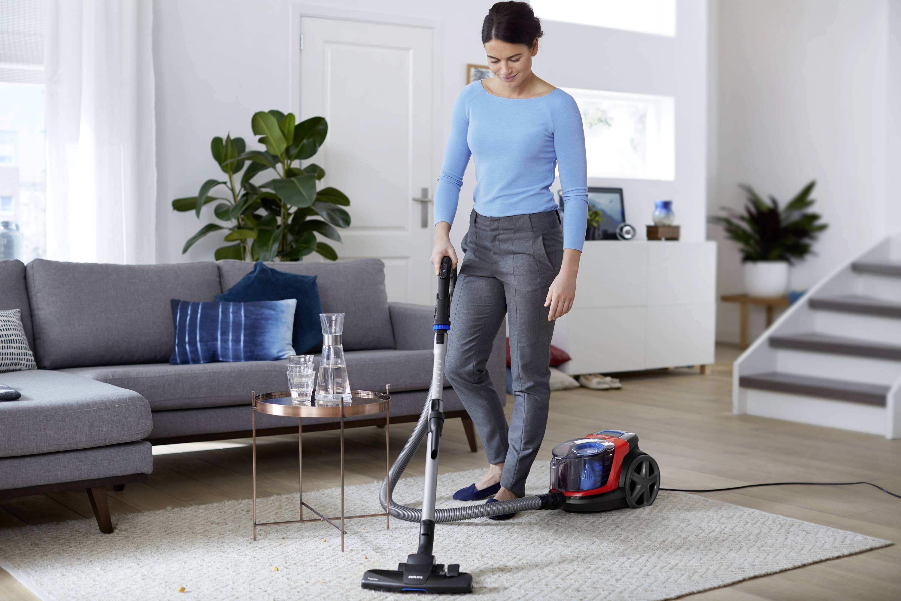 Eine Frau saugt mit einem Staubsauger im Wohnzimmer. Sie steht auf einem hellen Teppich, neben einem grauen Sofa und rundem Tisch.