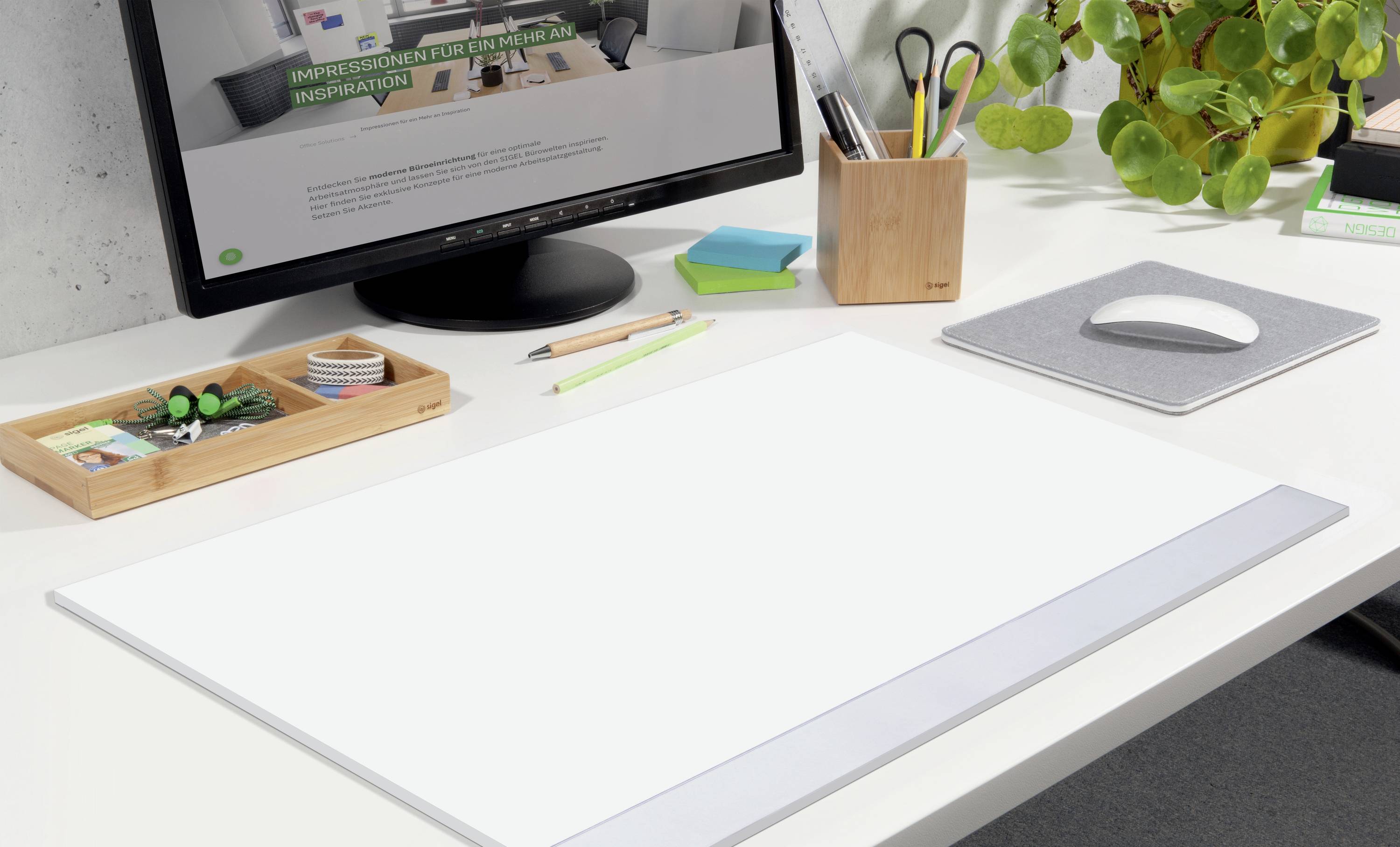 Modernes, aufgeräumtes Büro mit Monitor und Zubehör; Fokus auf weiße Schreibtischunterlage. Pflanzen und Schreibutensilien im Hintergrund.