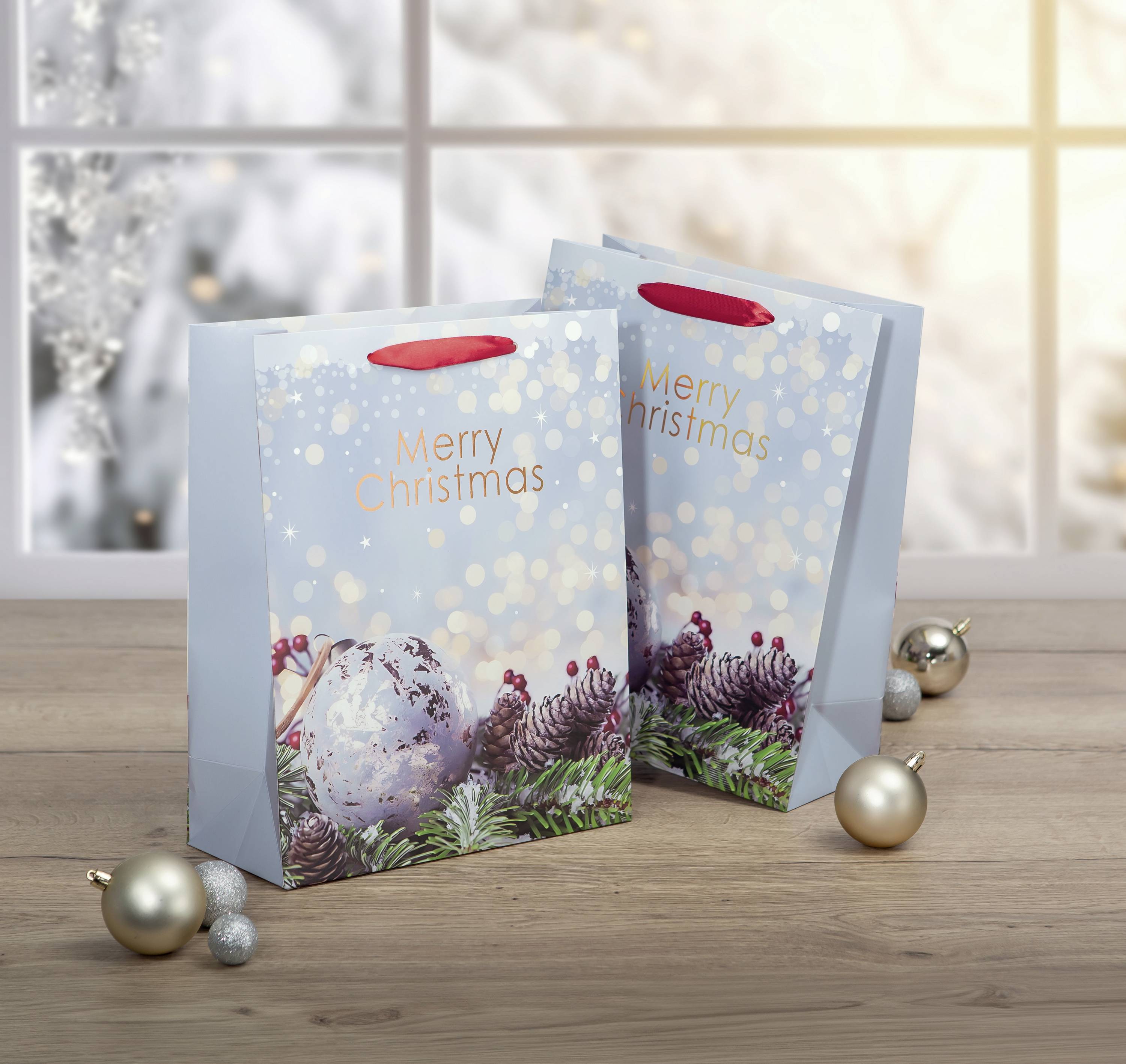 Zwei weihnachtlich dekorierte Geschenktüten mit der Aufschrift 'Merry Christmas' stehen auf einem Holztisch, umgeben von silbernen Kugeln.