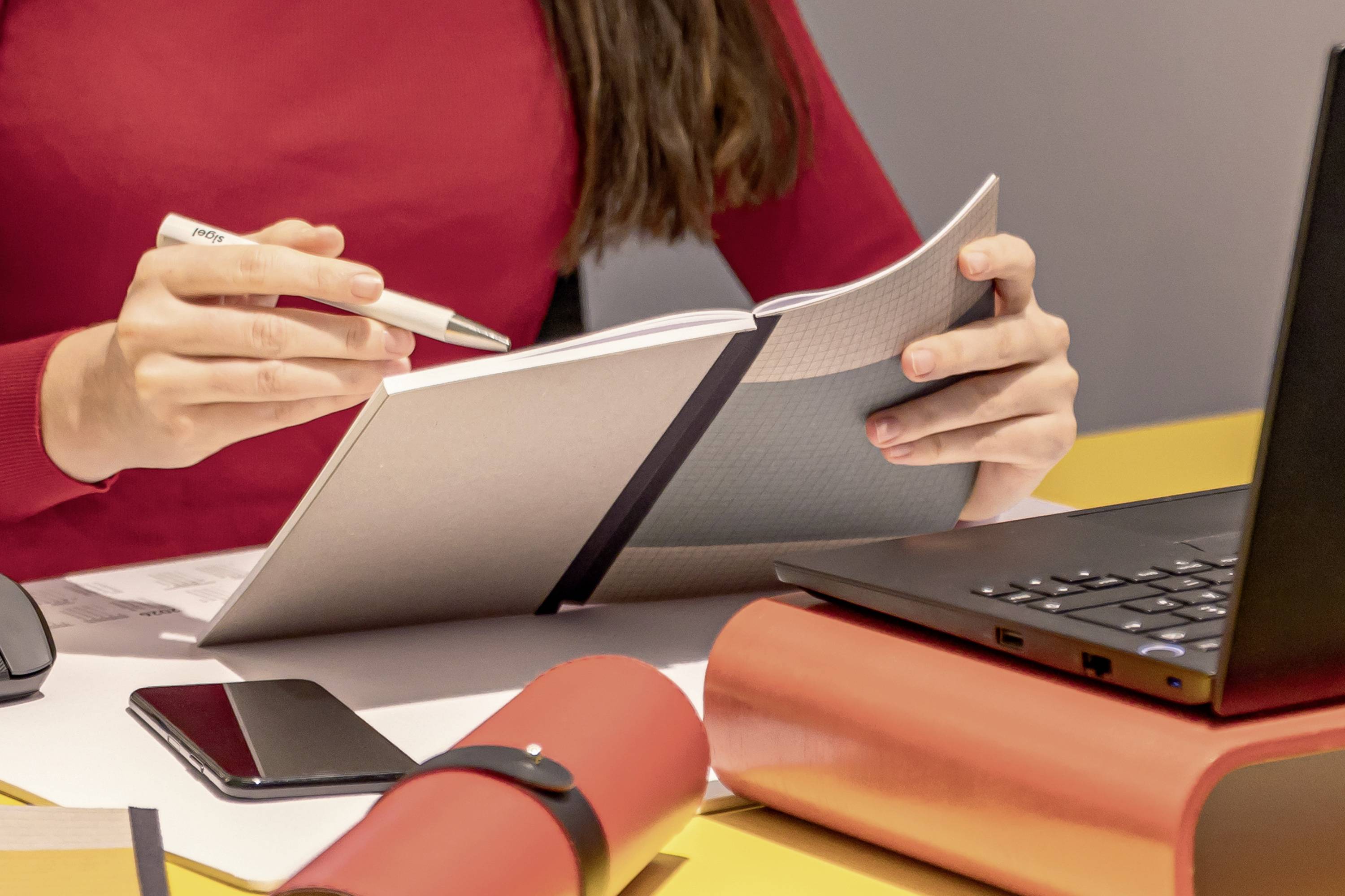 Eine Person in rotem Pulli schreibt mit einem Stift in ein Notizbuch. Auf dem Tisch liegen ein Laptop, ein Smartphone und andere Büroartikel.